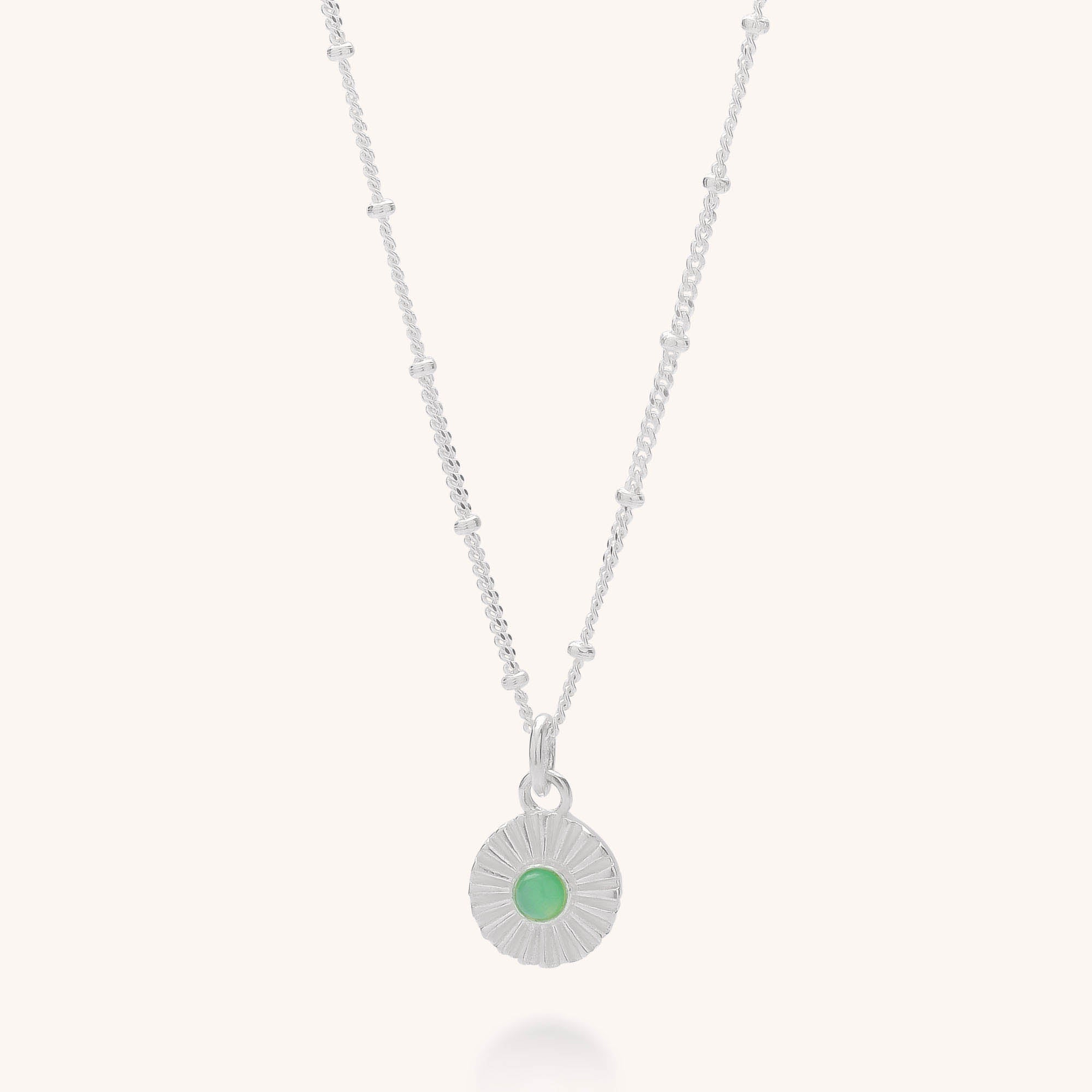 The Radiance Birthstone Necklace - May features a silver sunburst-patterned round pendant with a green chrysoprase birthstone, set on a delicate beaded chain and displayed against a light background.