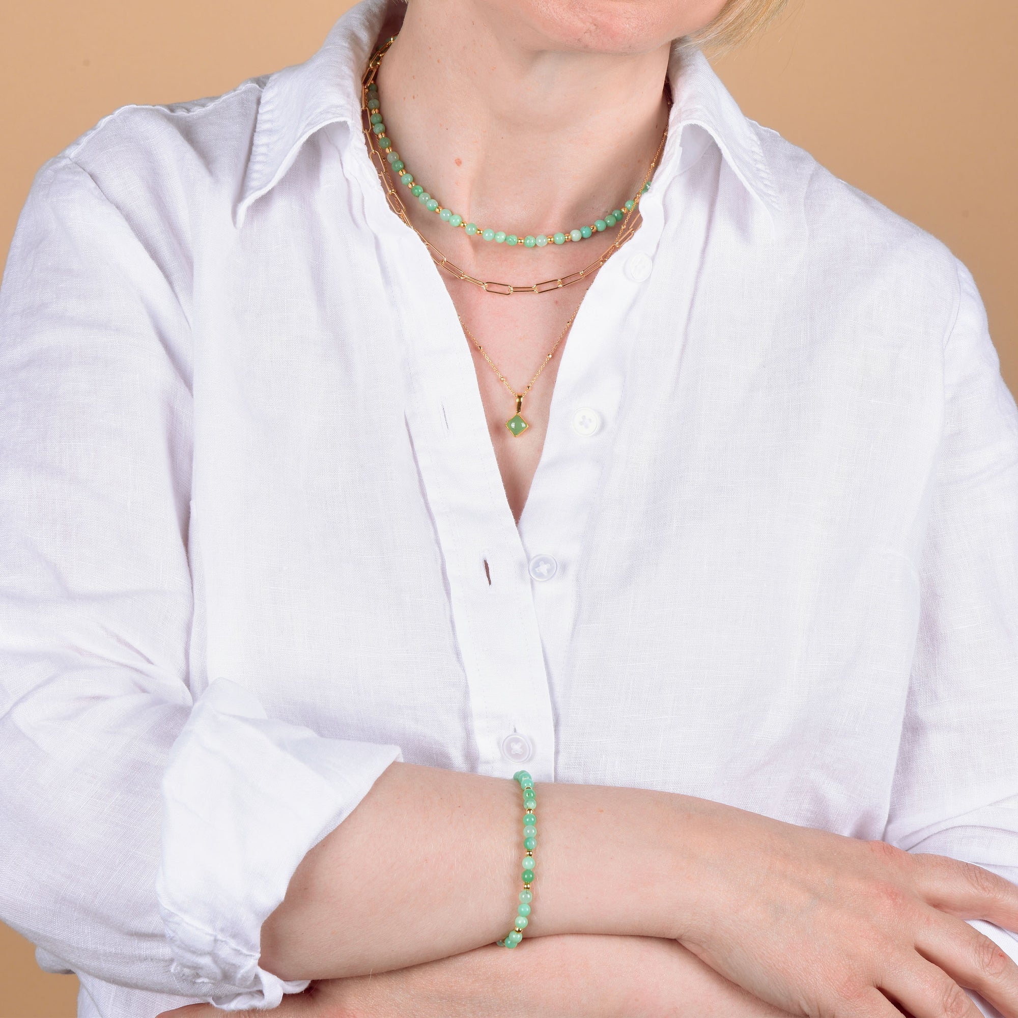 A person in a white shirt wears layered gold necklaces with green beads and the Jewel Bead Birthstone Bracelet - May, posing with arms crossed against a beige background.