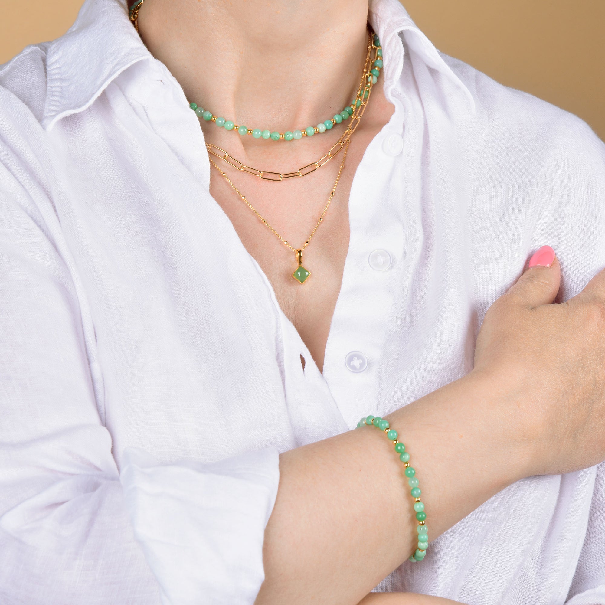 A person in a white button-down shirt showcases layered gold and green gemstone necklaces, the Jewel Bead Birthstone Bracelet - May, and pink nail polish on one hand, all set against a beige background.