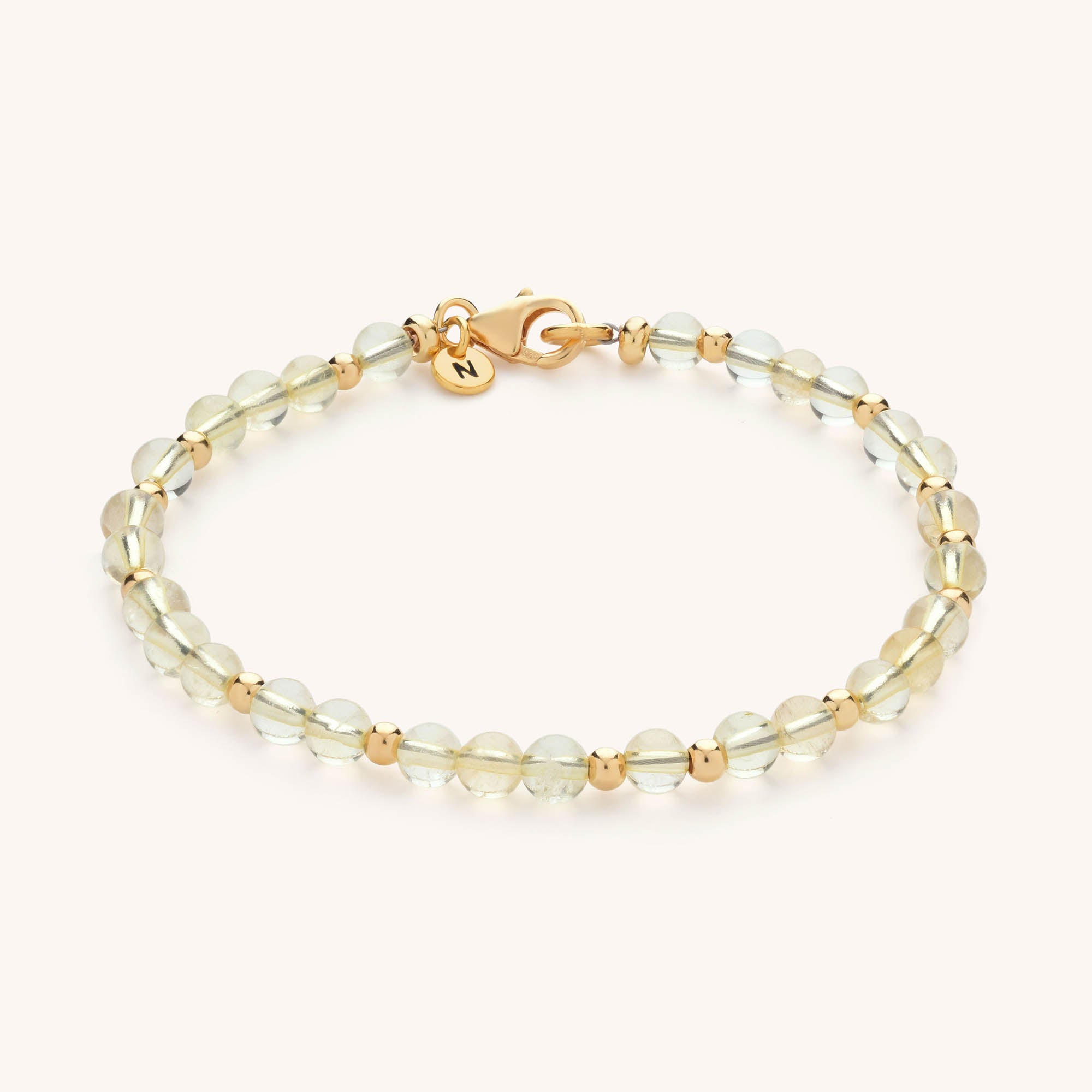 The Jewel Bead Birthstone Bracelet features translucent round beads, small gold spacers, a gold clasp, and a round gold charm engraved with “N.”.