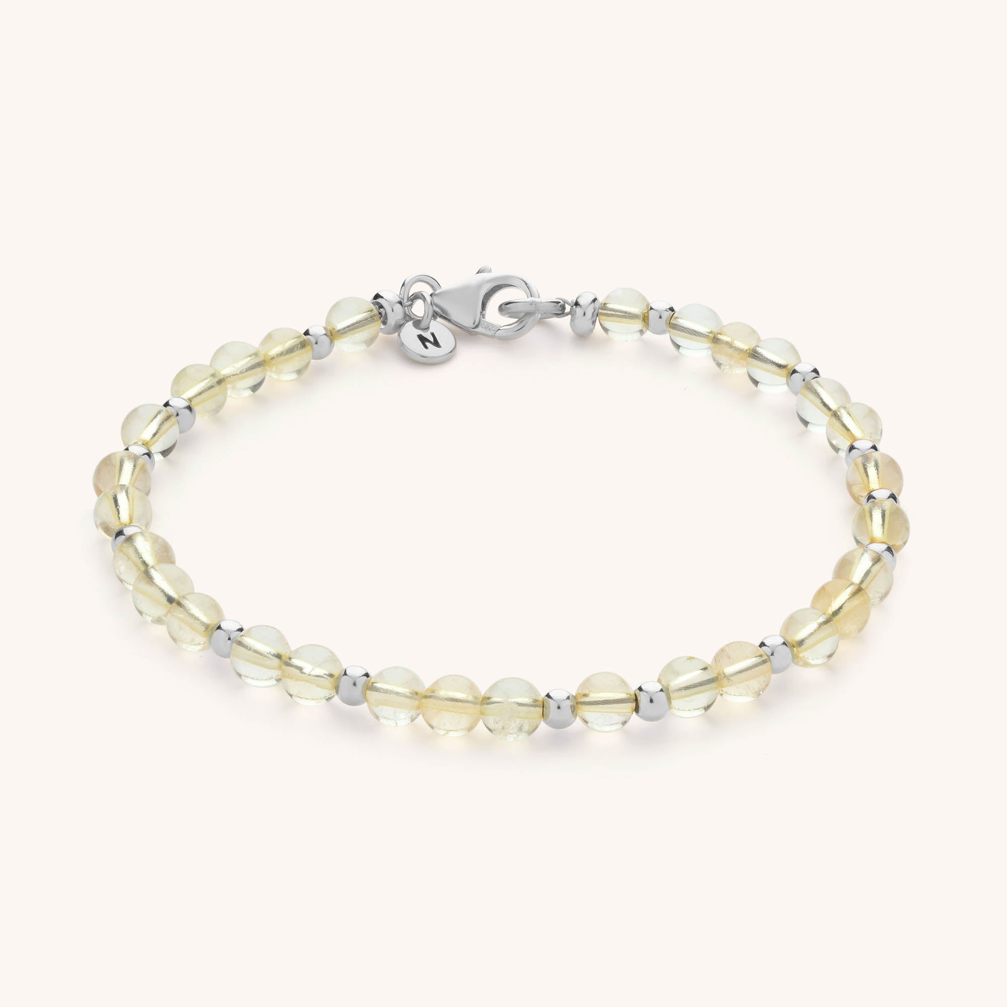 The Jewel Bead Birthstone Bracelet features round, pale yellow beads alternating with small silver beads, finished with a silver lobster clasp and a dainty round charm—a beautifully handmade, elegant accessory.