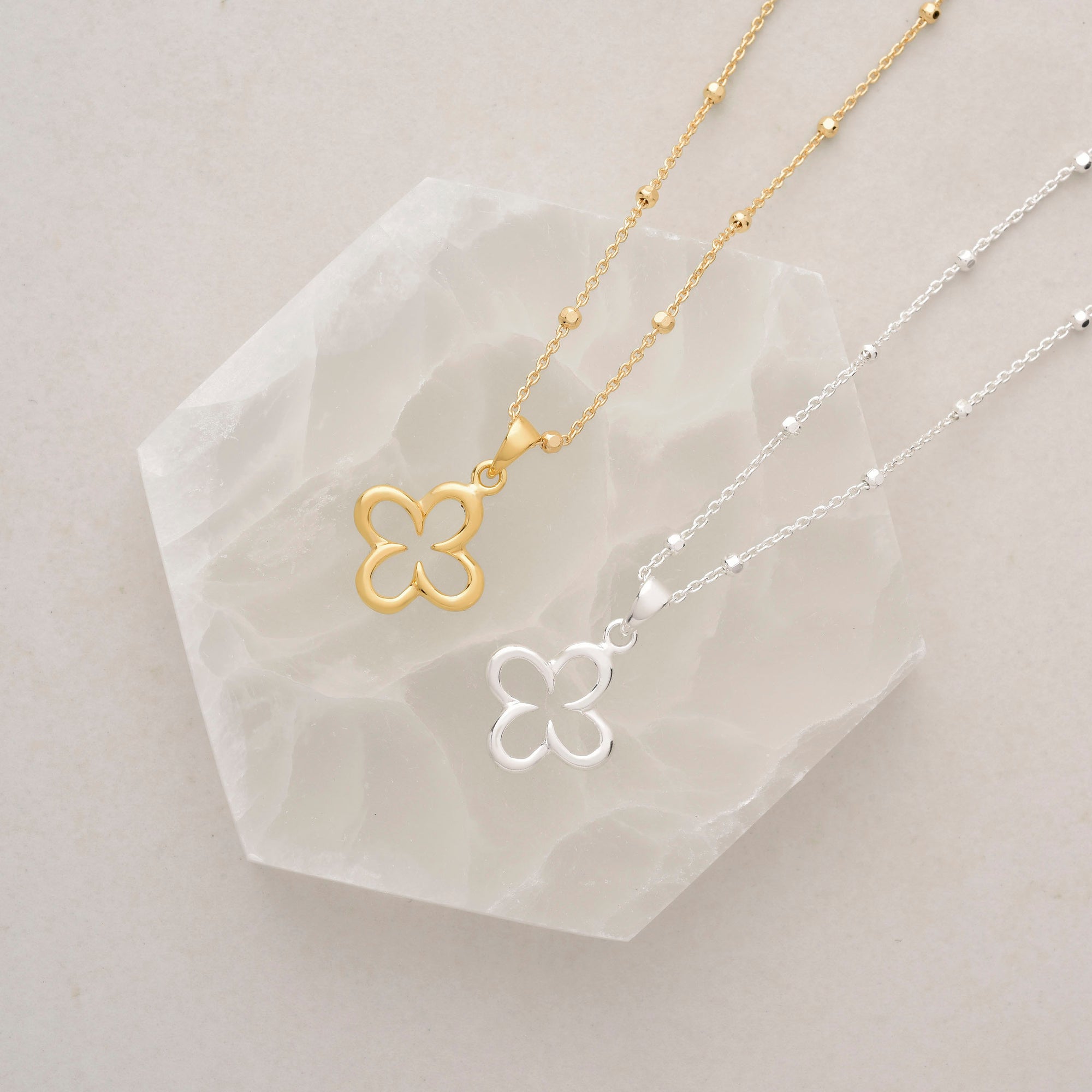 The Clover Necklace is shown in both gold and silver on a white, hexagonal stone slab with a light beige background—a charming addition to any jewellery collection.
