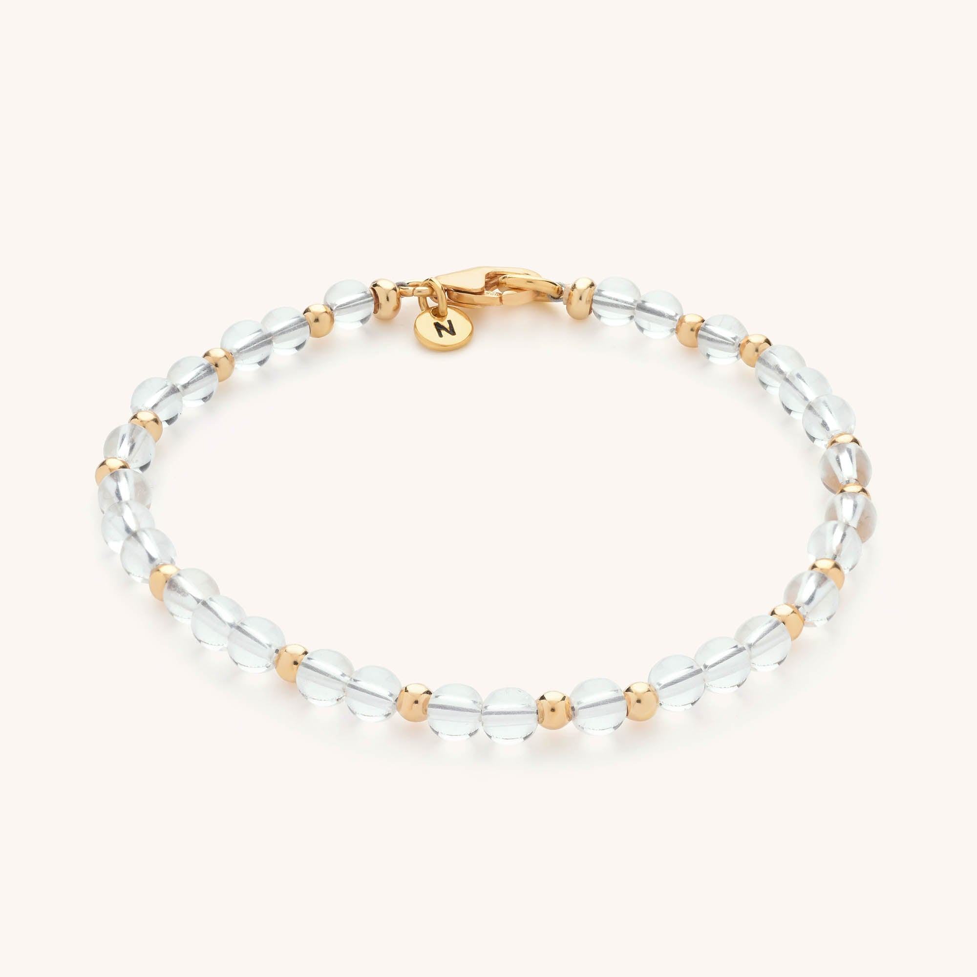 The Jewel Bead Birthstone Bracelet features alternating clear round beads and small gold beads, a gold clasp, and a round gold N charm—a perfect handmade piece for jewellery lovers.