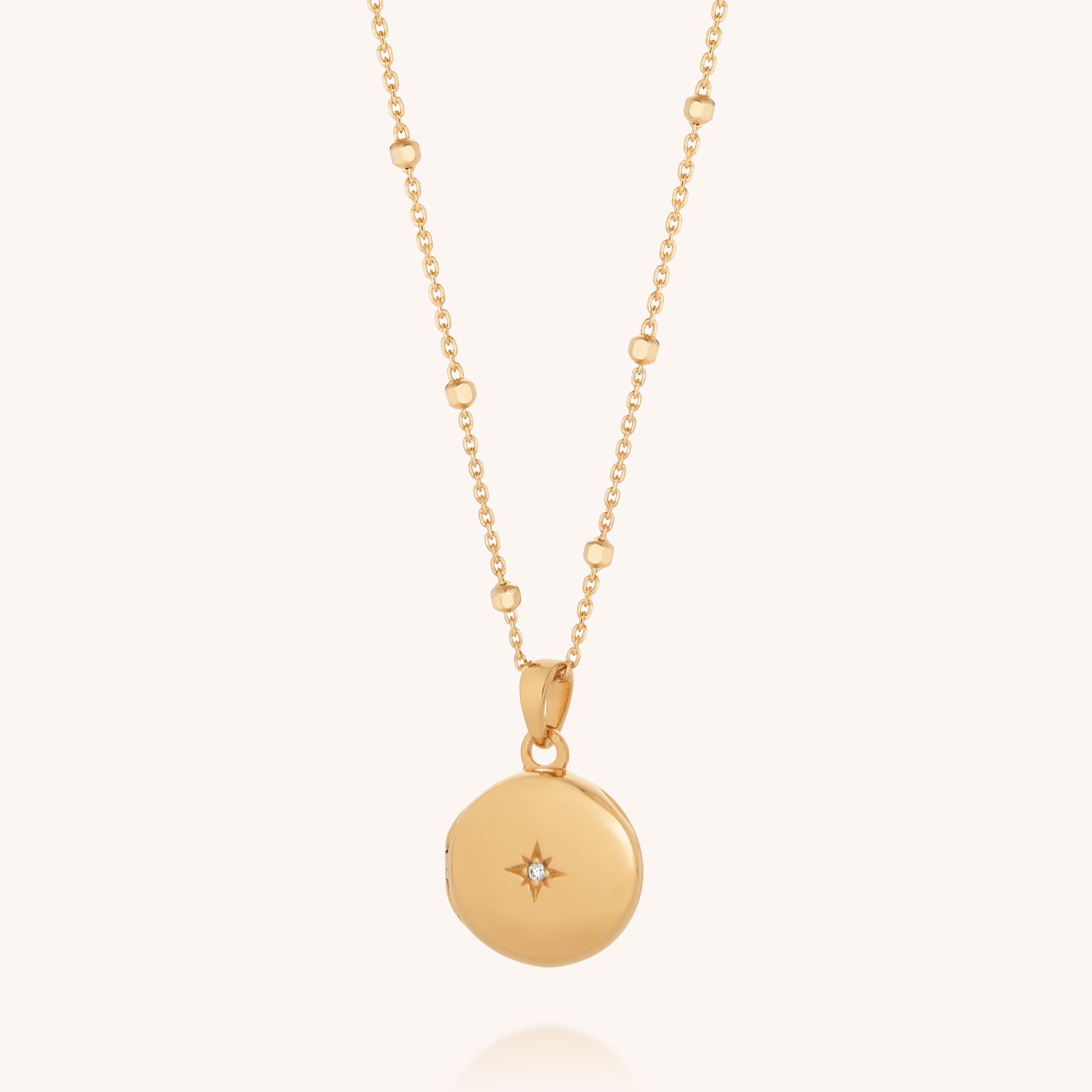The Starlight Keepsake Locket Necklace features a round gold pendant with a central gemstone and a delicate chain accented by spaced gold beads, creating an elegant piece of celestial-inspired jewellery.