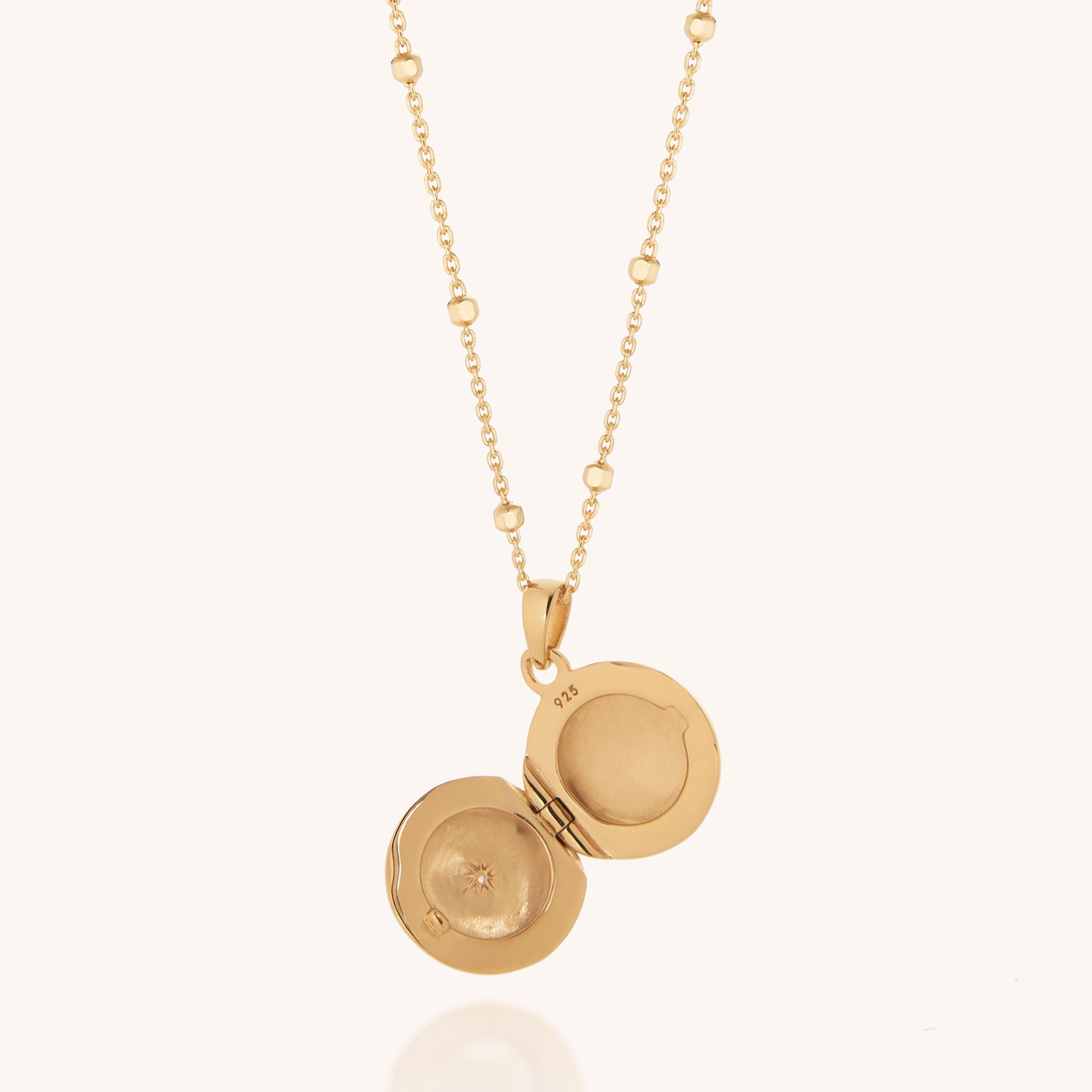 The Starlight Keepsake Locket Necklace features a gold circular hinged pendant with an empty interior. The chain includes small accent beads, and its celestial charm stands out against the plain, light background.