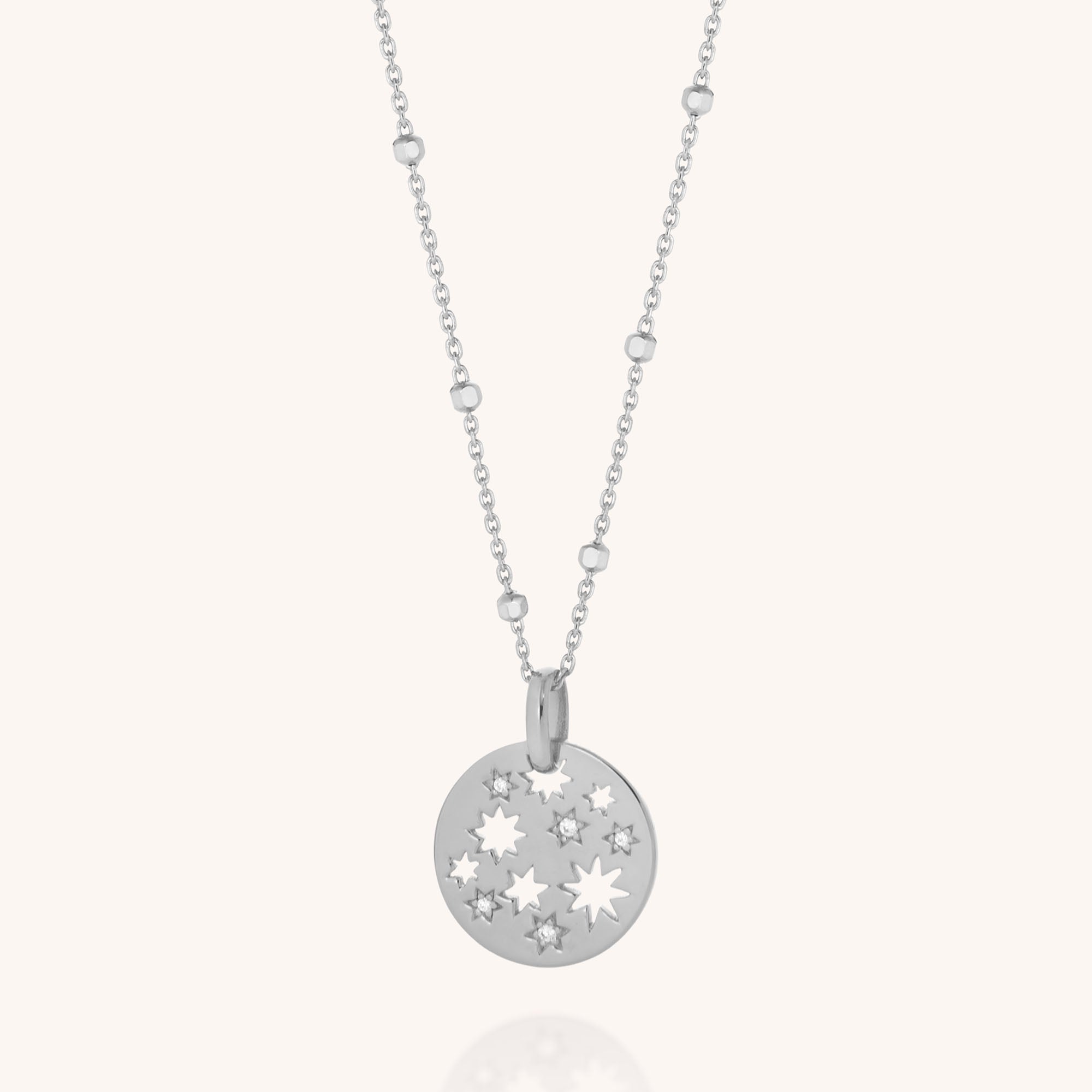 The Starry Skies Necklace features a silver round pendant with cut-out star shapes and cubic zirconia accents on a beaded chain, shown against a plain light background.