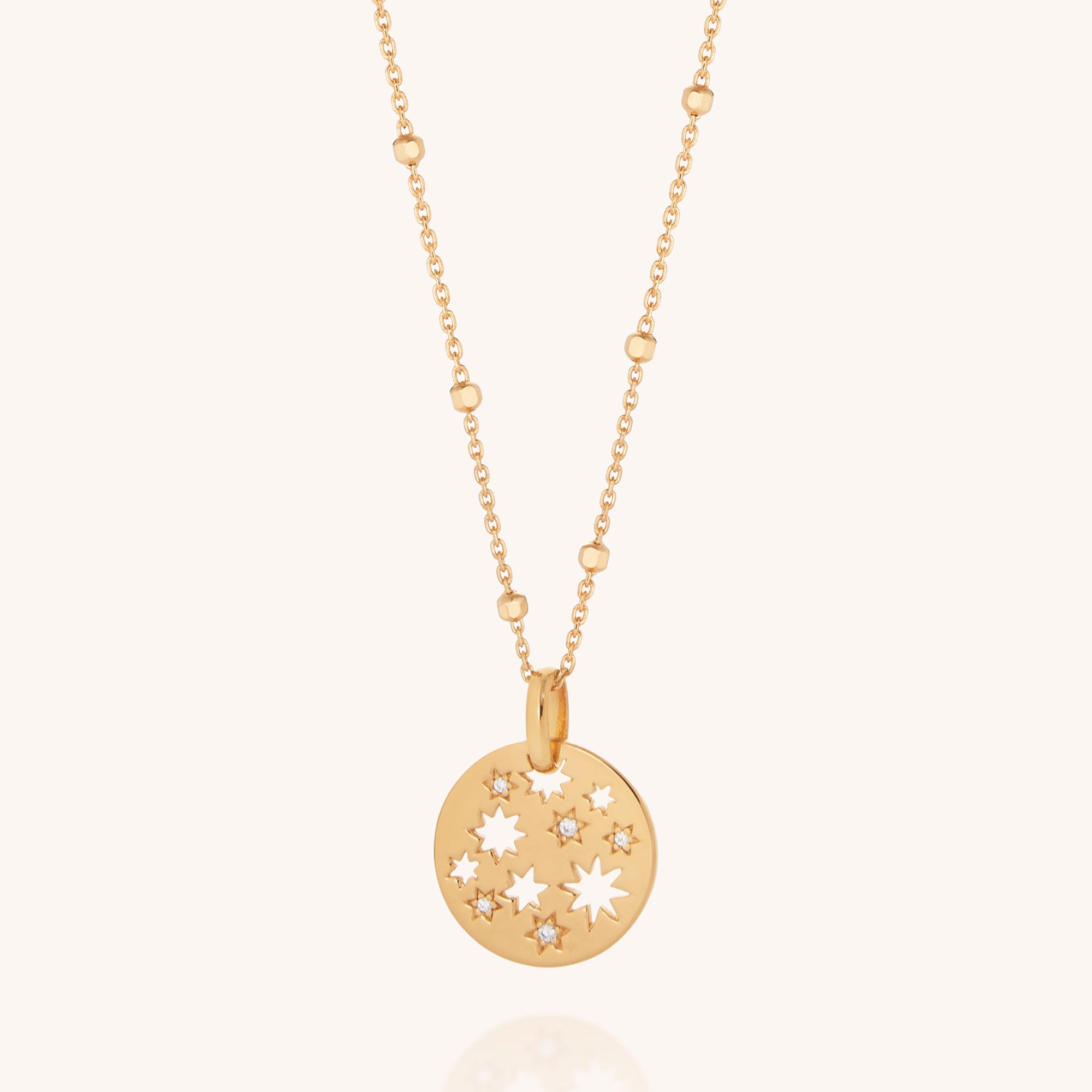 The Starry Skies Necklace features a gold chain with small, evenly spaced beads and a round pendant with star cutouts and cubic zirconia accents, set against a plain light background.