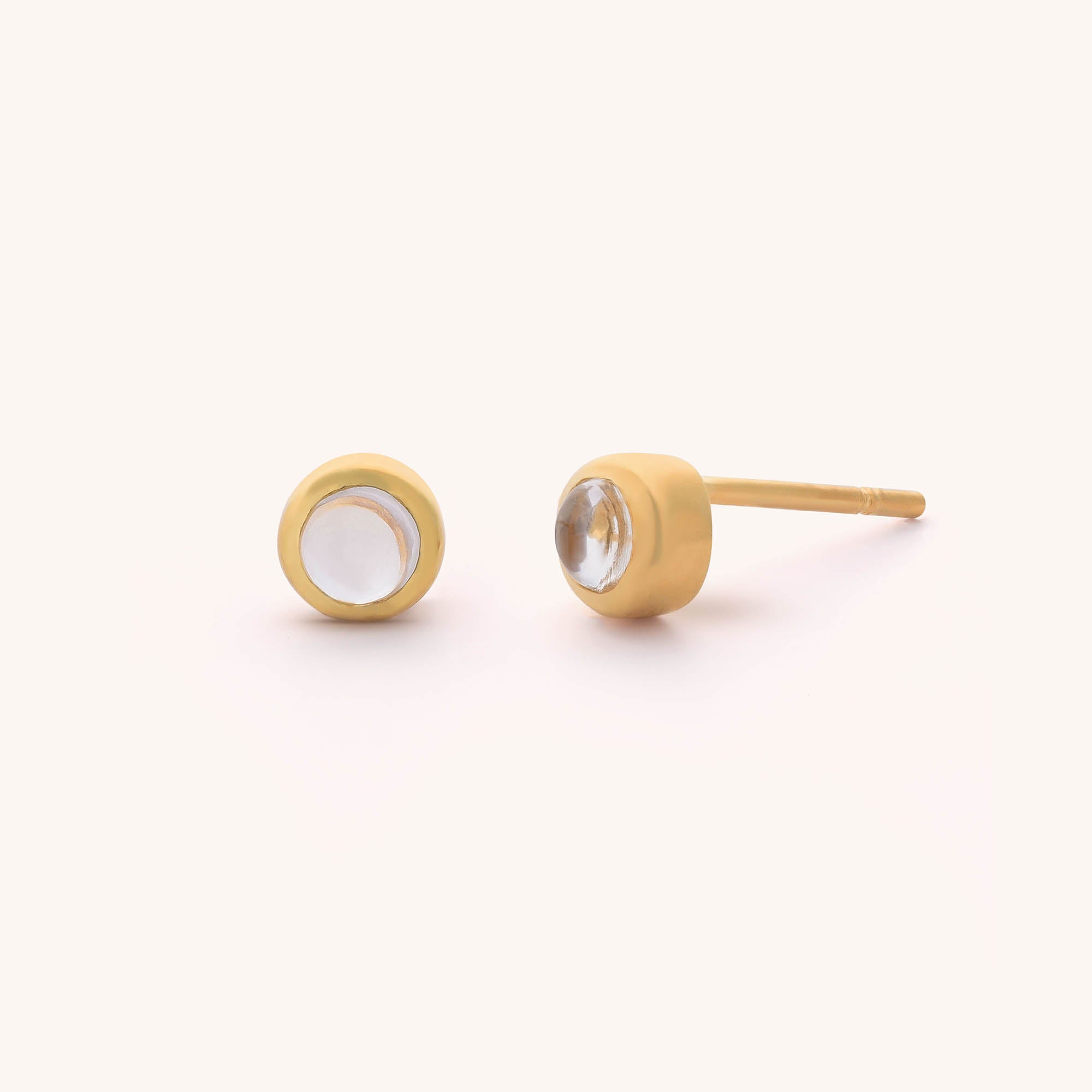 The Mini Gem Dot Birthstone Earrings for April feature round, clear gemstone centers in gold settings. One earring stands upright to highlight the gem, while the other lies on its side, showcasing the post and setting against a warm light beige background.