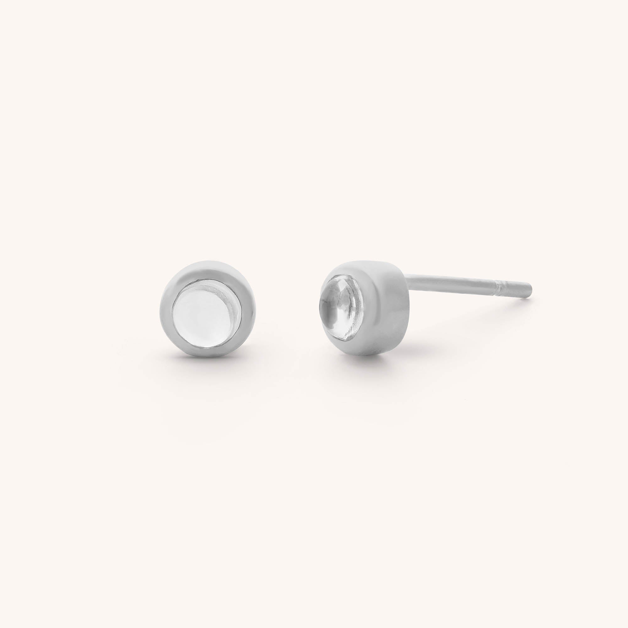 The Mini Gem Dot Birthstone Earrings - April feature two minimalist sterling silver stud earrings with circular settings and clear crystal quartz. One earring is upright while the other lies on its side, all set against a plain white background for an elegant touch.