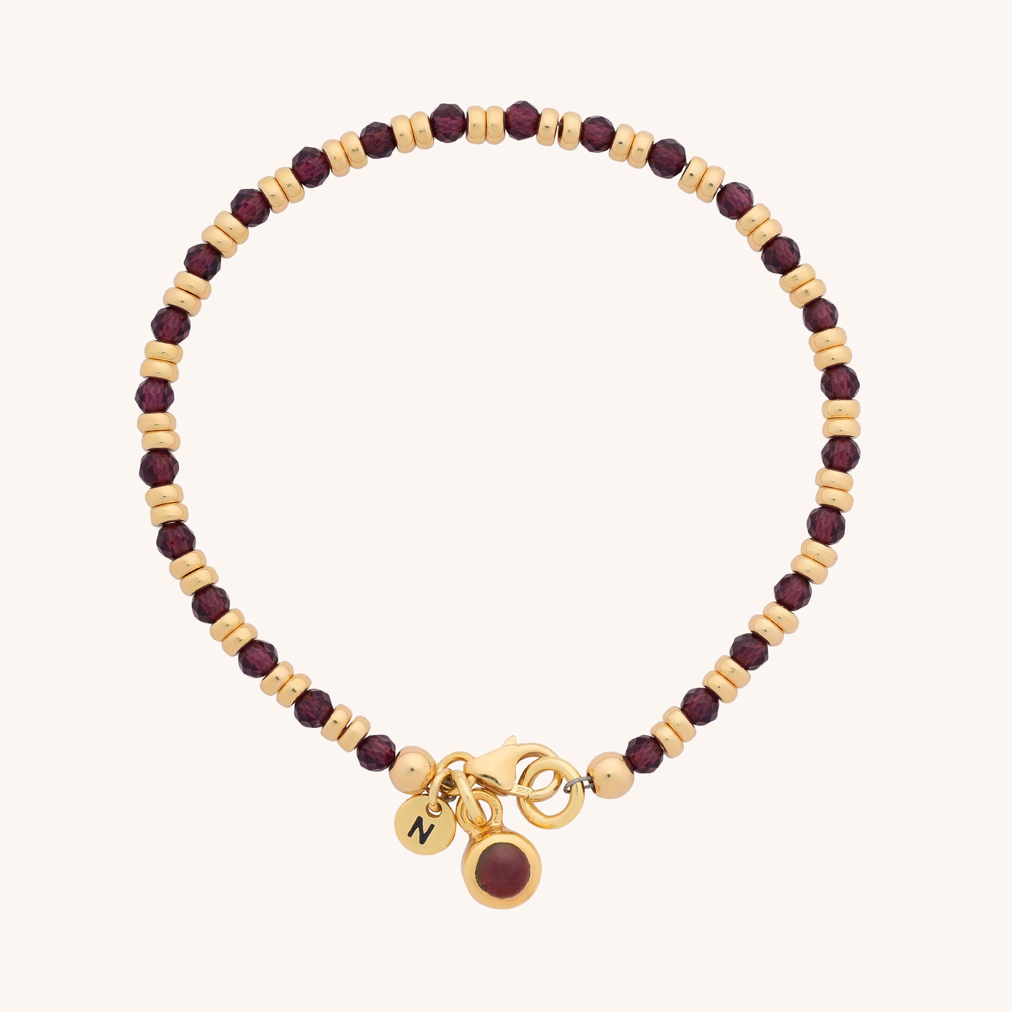 The Linear Birthstone Bracelet - January features alternating small gold and dark red beads, with three gold charms: a round red gem and a disc engraved with the letter Z.