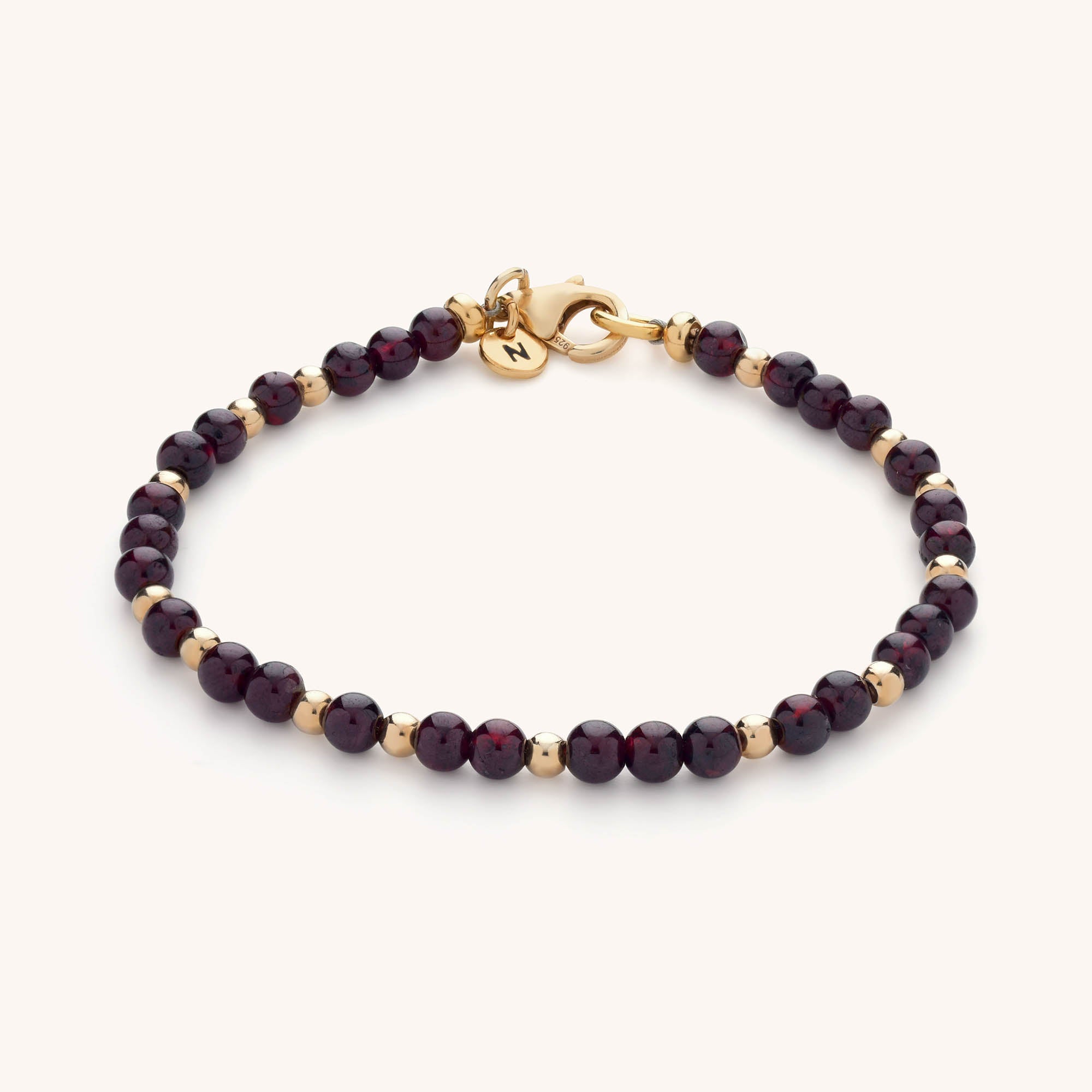 The Jewel Bead Birthstone Bracelet features alternating small round dark red and gold beads, a gold clasp, and a tiny gold charm engraved with the letter N—an elegant piece of handmade jewellery.