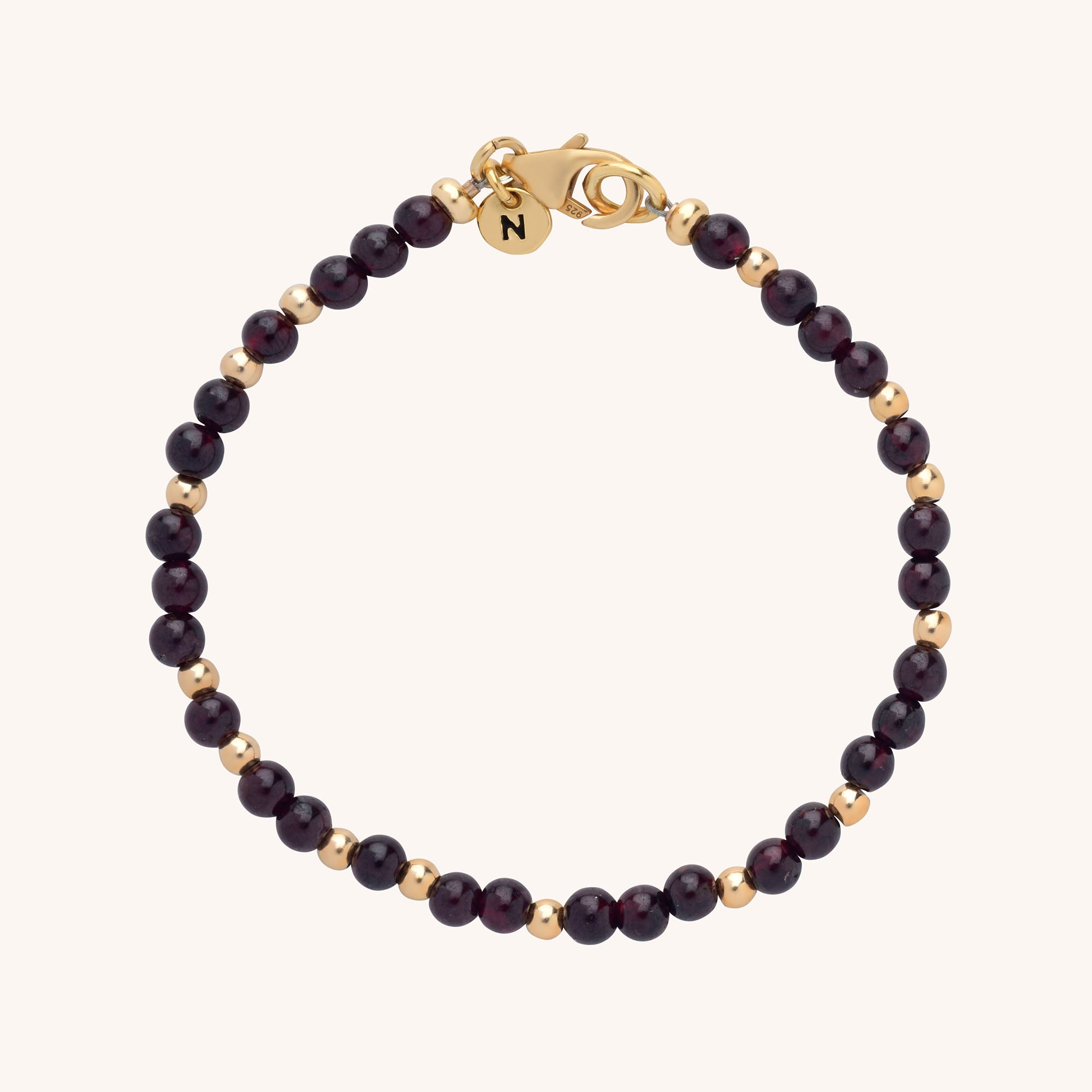 The Jewel Bead Birthstone Bracelet features alternating small gold and dark red beads, finished with a gold lobster clasp and a round gold charm engraved with the letter “N”—a perfect handmade jewellery piece.