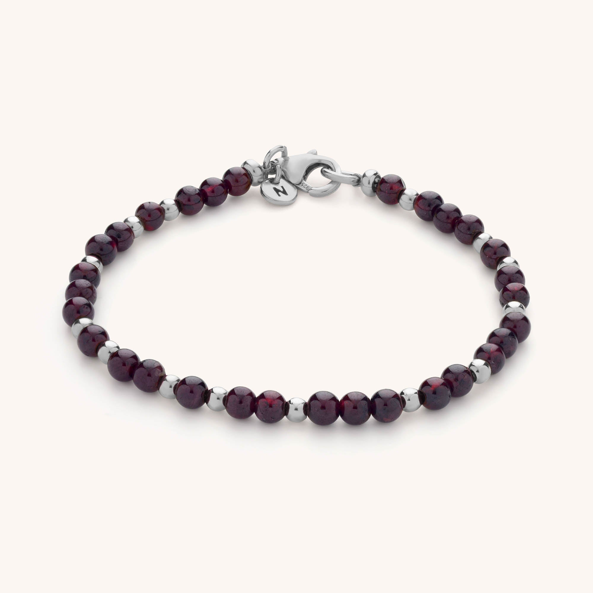 The Jewel Bead Birthstone Bracelet features alternating deep red and silver round beads, a secure silver clasp, and a charm engraved with the letter N—an elegant piece of handmade jewellery.