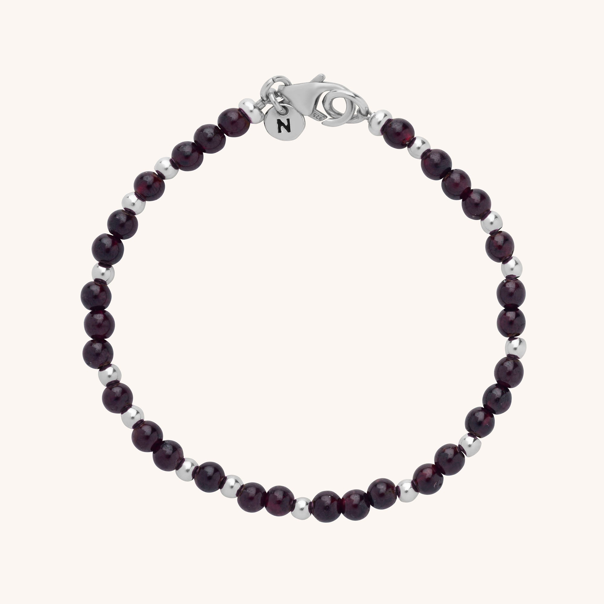 A delicate handmade Jewel Bead Birthstone Bracelet features alternating small dark red and silver beads, a silver lobster clasp, and a round charm engraved with the letter N. Displayed on a light background.