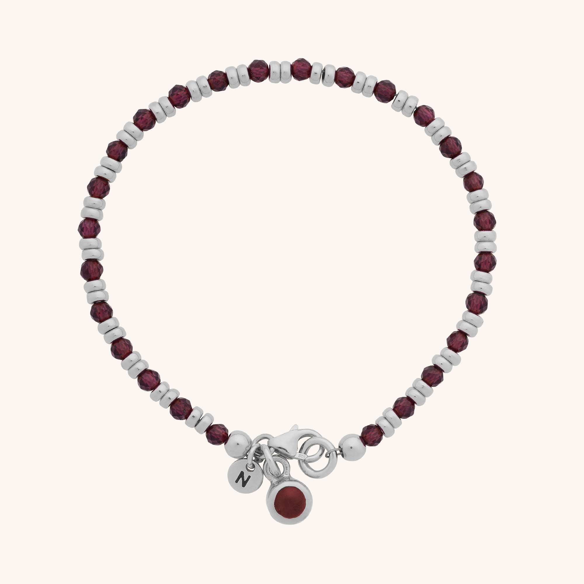 The Linear Birthstone Bracelet - January features alternating silver and dark red beads, a clasp, and two small dangling charms—one engraved with an N and another adorned with a sparkling garnet gemstone.