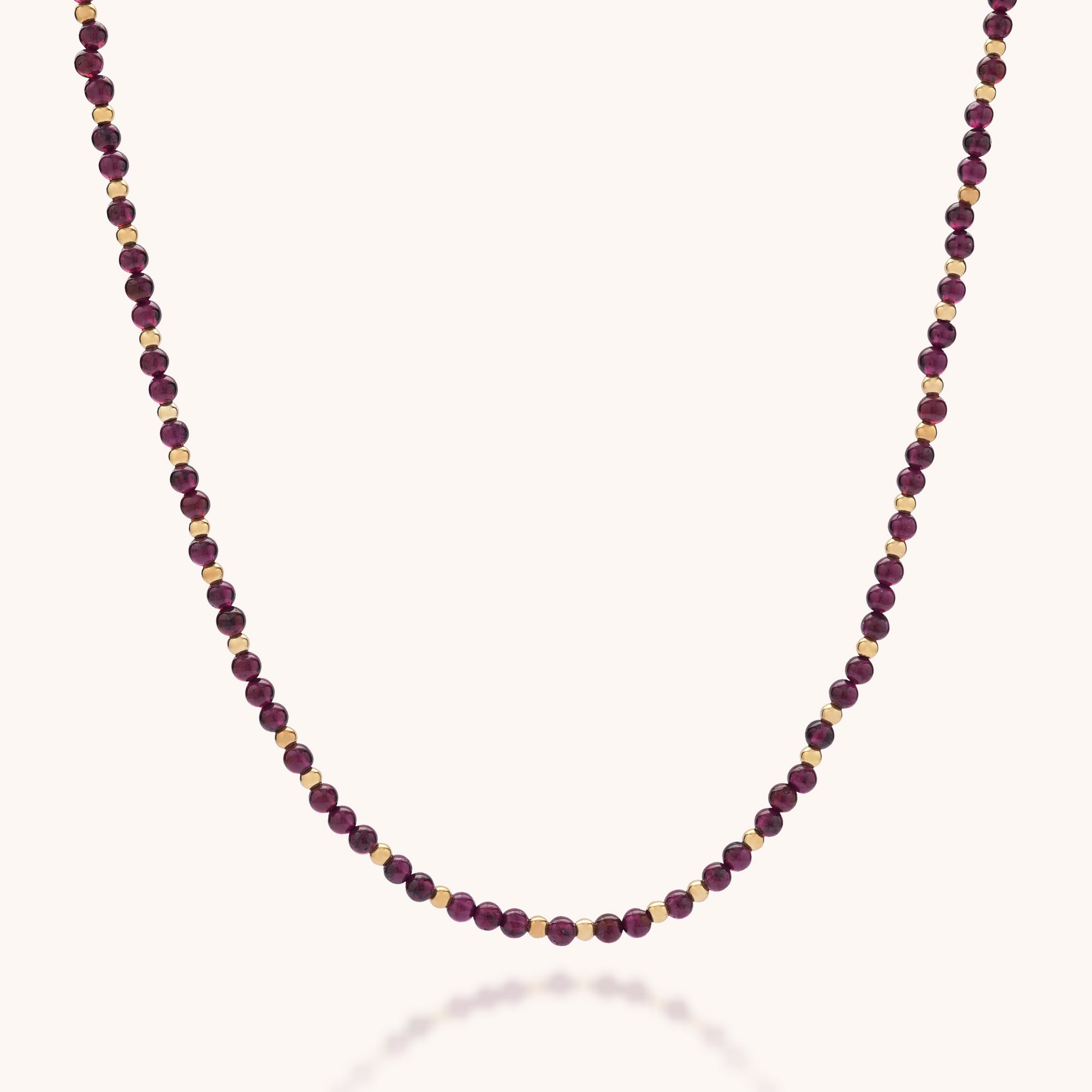 The Jewel Bead Birthstone Necklace - January features handmade alternating small round gold and deep red garnet beads in a simple, elegant pattern against a light background.
