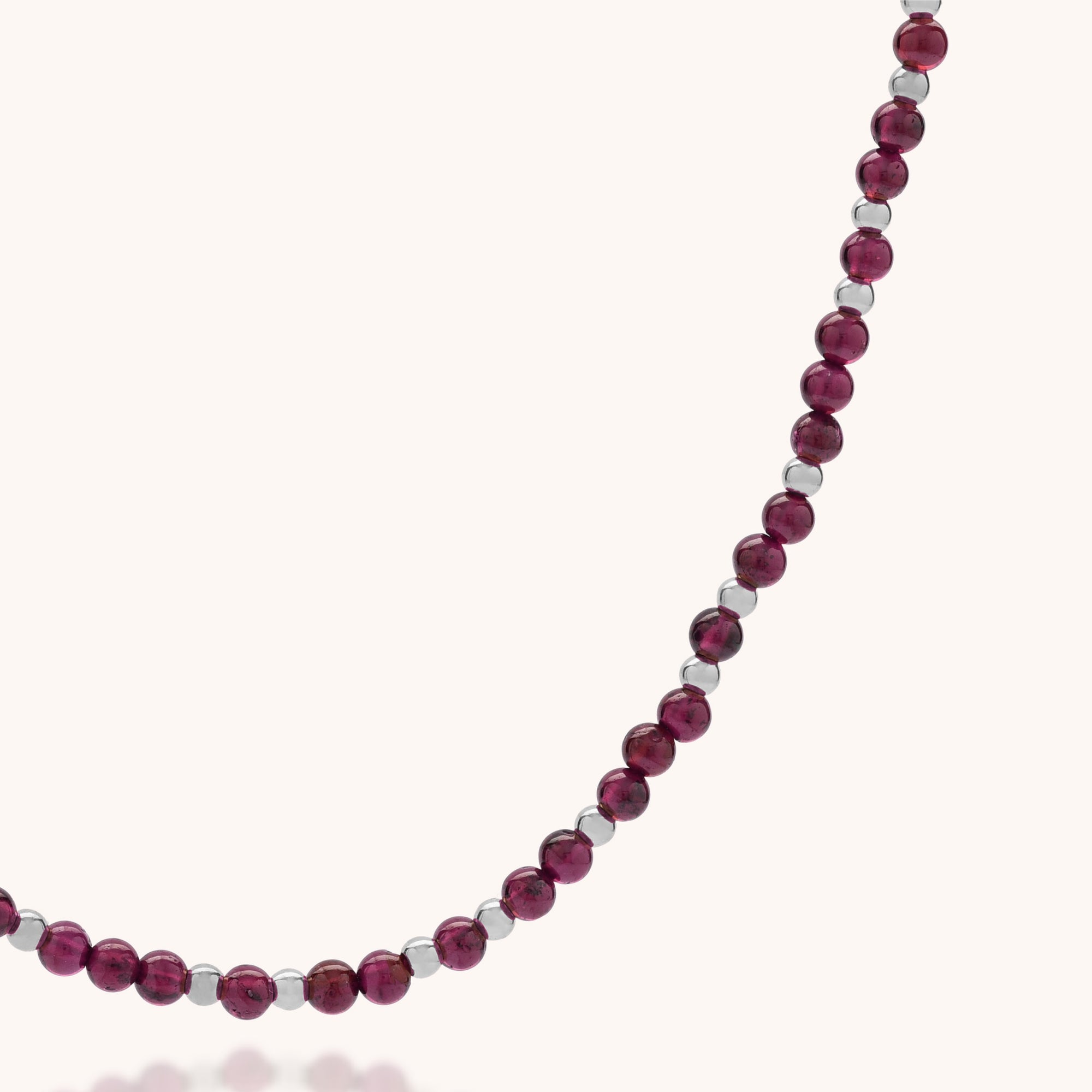 A close-up of the Jewel Bead Birthstone Necklace - January, showcasing alternating shiny silver and deep red garnet beads arranged in a gentle curve on a plain, light background.