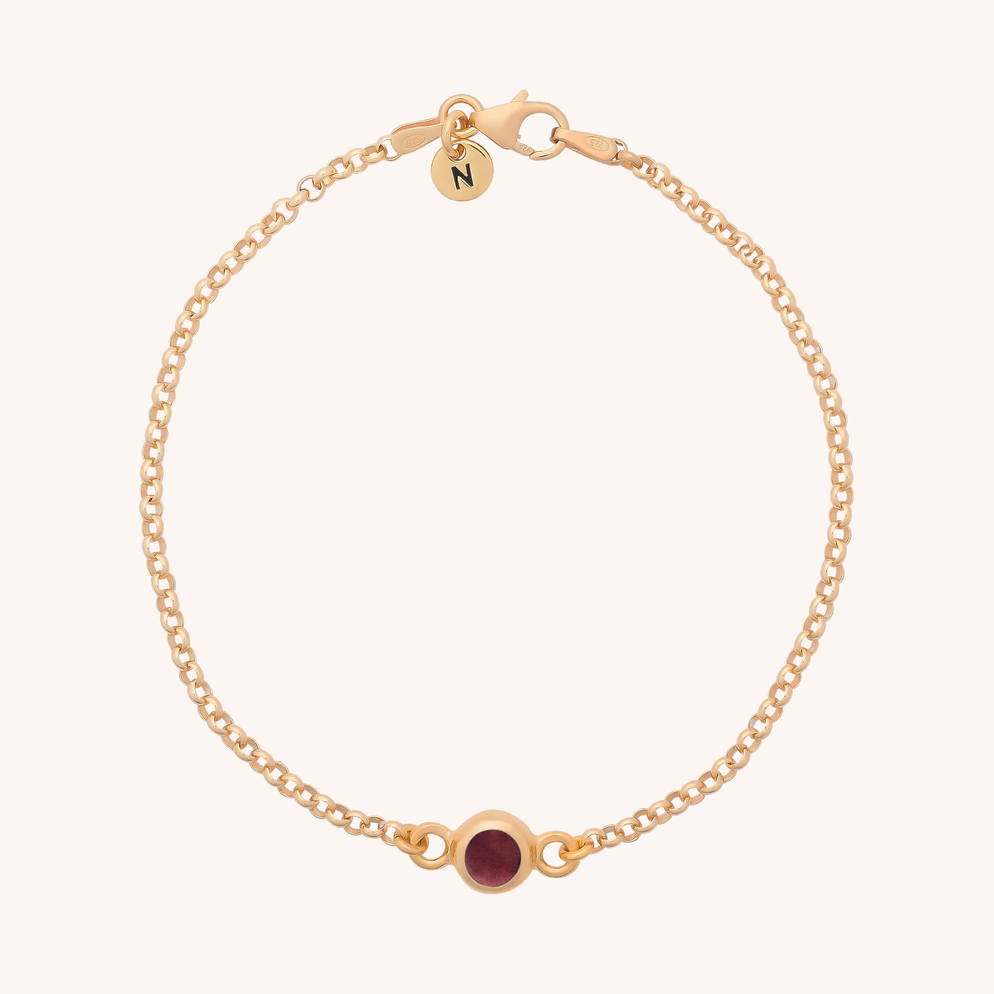 The Birthstone Gem Dot Bracelet features a delicate gold chain with a small round red gemstone in the center and a circular N charm—perfect for lovers of personalized jewelry.