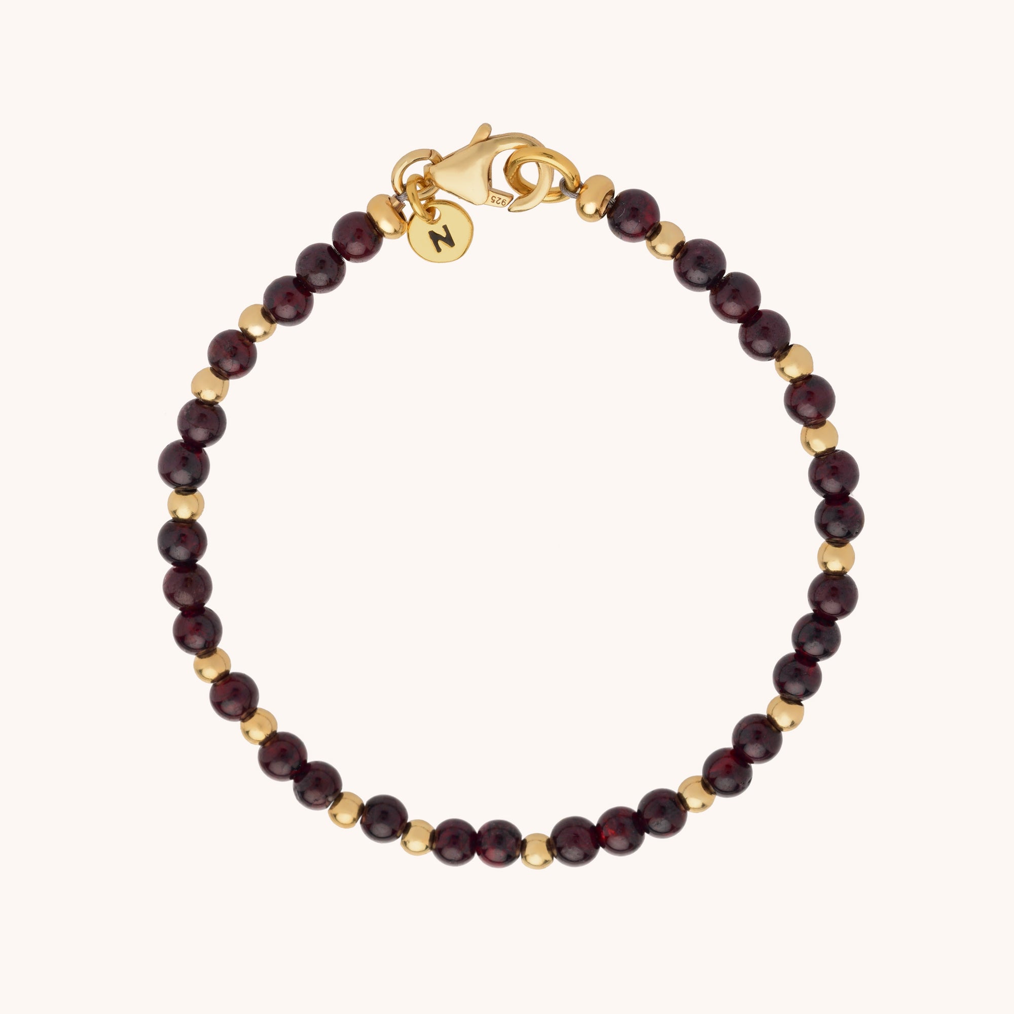 The Jewel Bead Bracelet features alternating dark red natural gemstone beads and small gold beads, finished with a gold lobster clasp and a round charm.