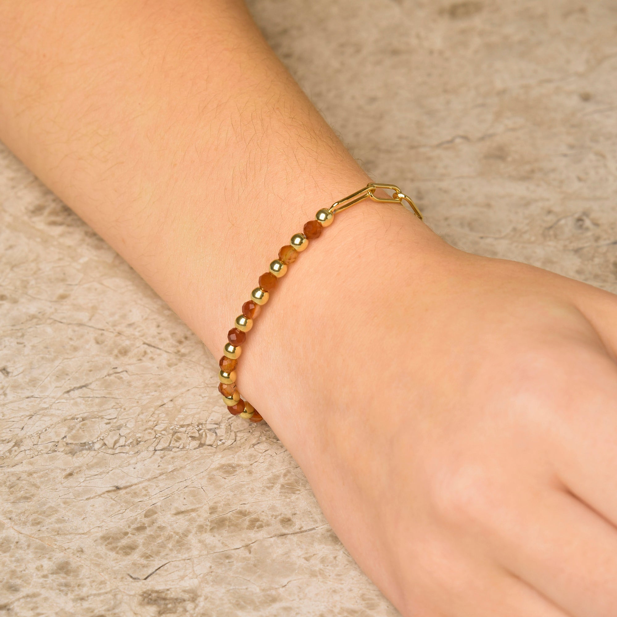 A close-up of a wrist adorned with the Gem Shine Bracelet, showcasing natural gemstone beads in brown and gold tones, set against a textured, light-colored background.