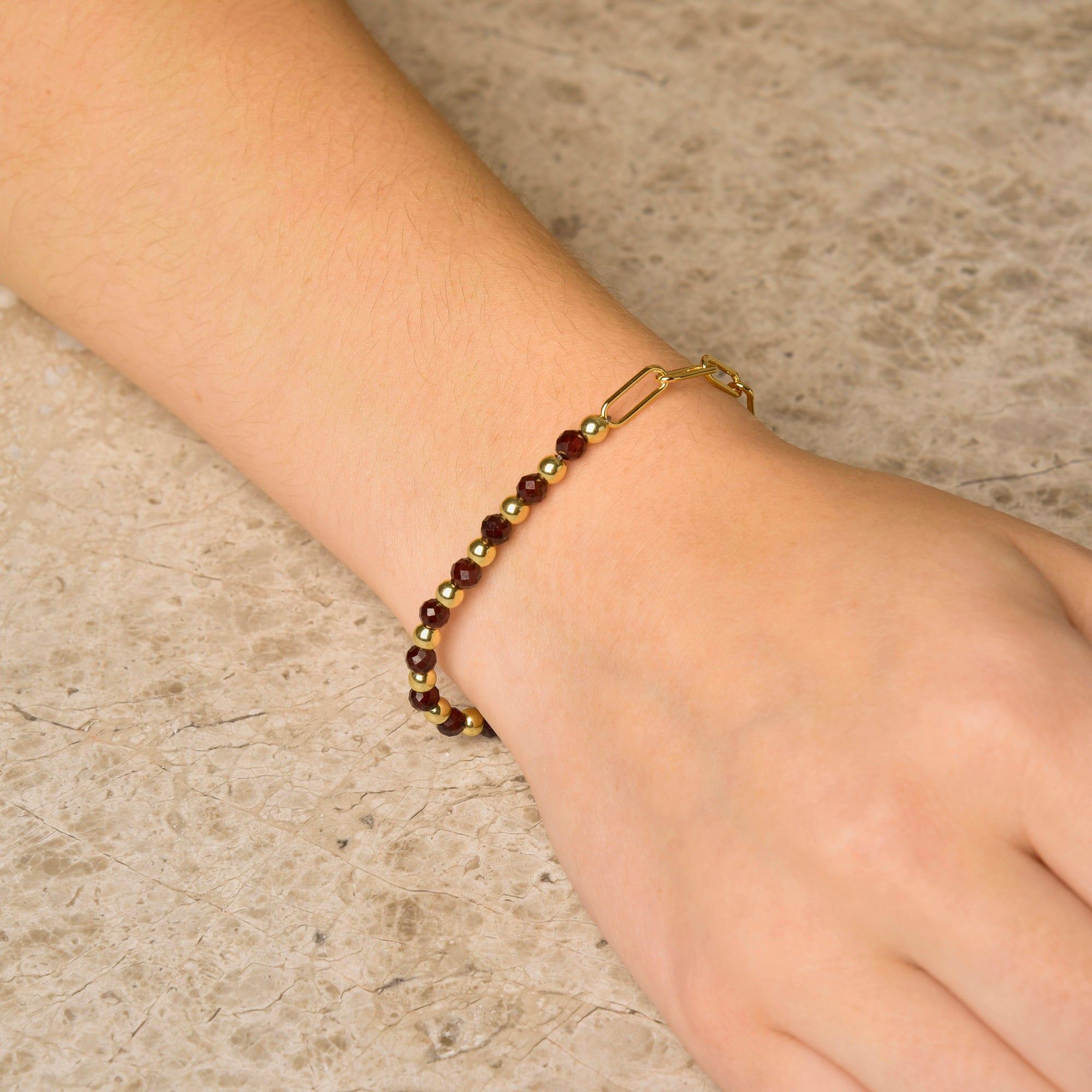 A hand rests on a textured stone surface, showcasing the Gem Shine Bracelet—featuring small, round red gemstones, gold accents, and a gold clasp.
