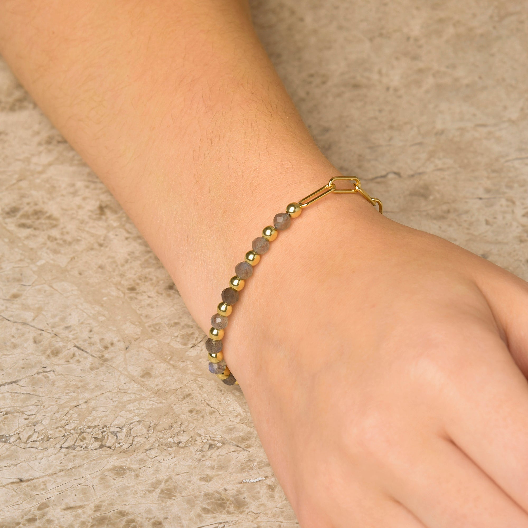 A hand rests on a light marble surface, wearing the Gem Shine Bracelet—delicate gray gemstones and gold links create an elegant look.