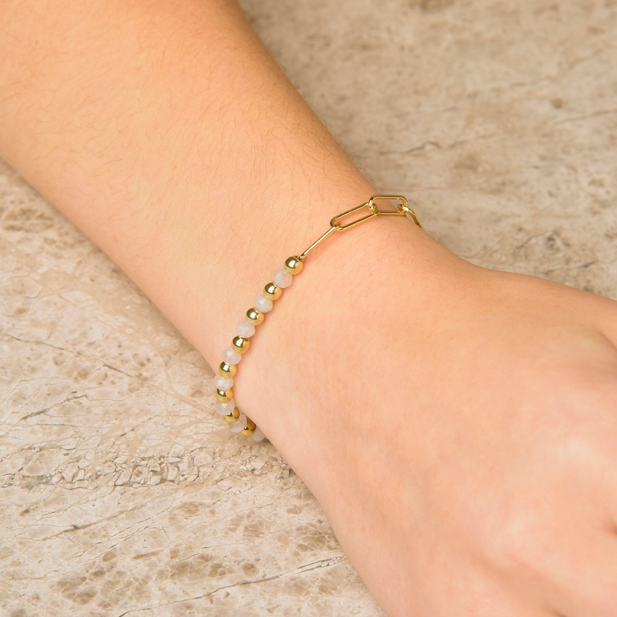 A close-up of a wrist wearing the Gem Shine Bracelet, adorned with small white and gold beads, natural gemstones, and a gold chain and clasp, resting on a light textured surface.