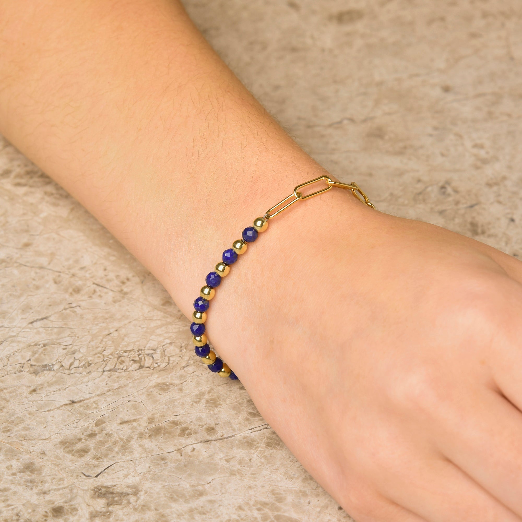 A hand rests on a textured stone surface, adorned with the Gem Shine Bracelet featuring small round blue natural gemstones and a gold paperclip-style chain.