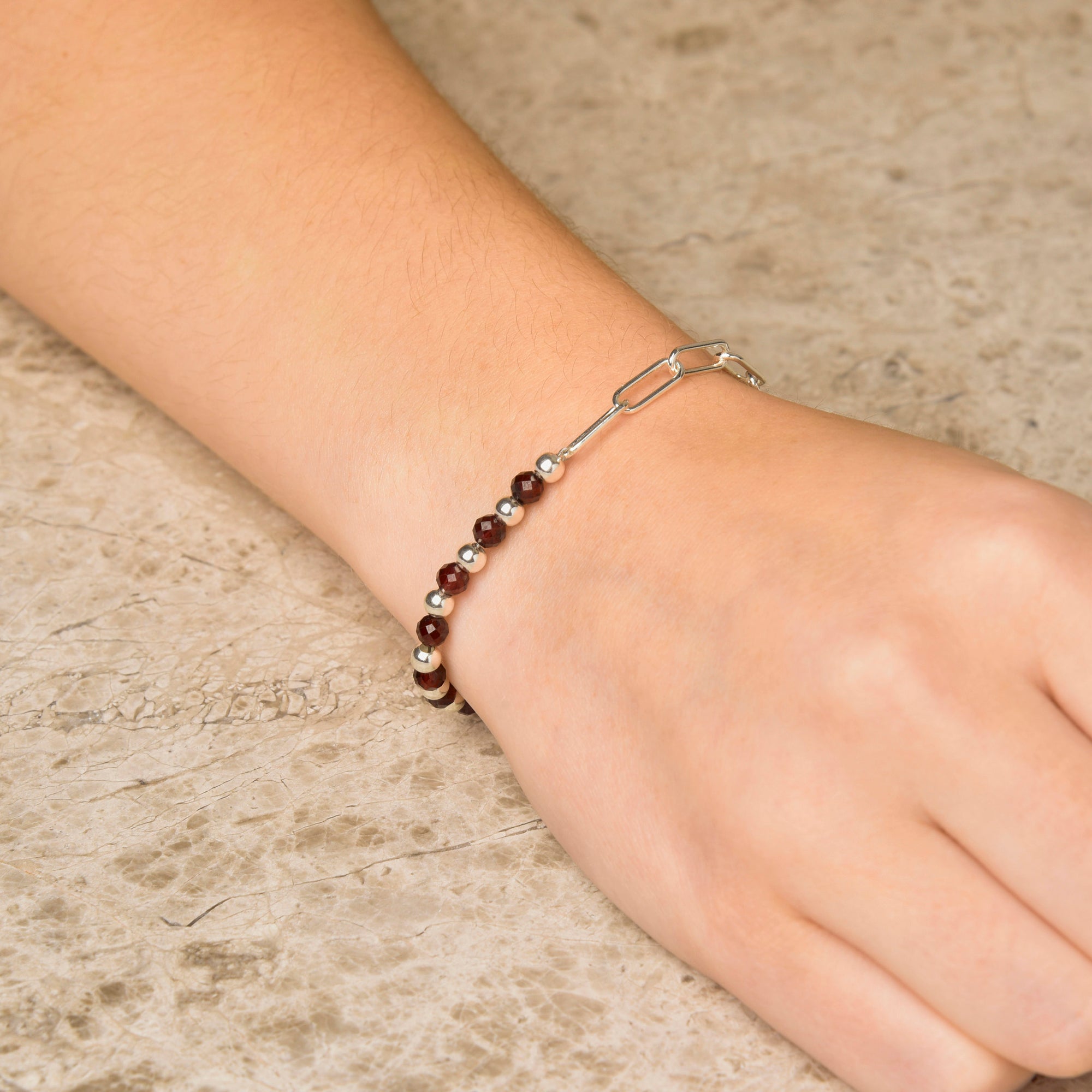 A hand rests on stone, adorned with the Gem Shine Bracelet—crafted from small dark red natural gemstones and eco-silver sterling silver links.