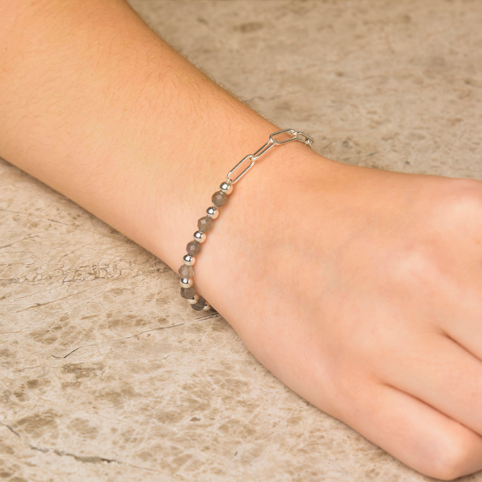 A close-up of a hand on textured stone, wearing the Gem Shine Bracelet—delicate chains and small metallic beads made from eco-silver sterling silver.