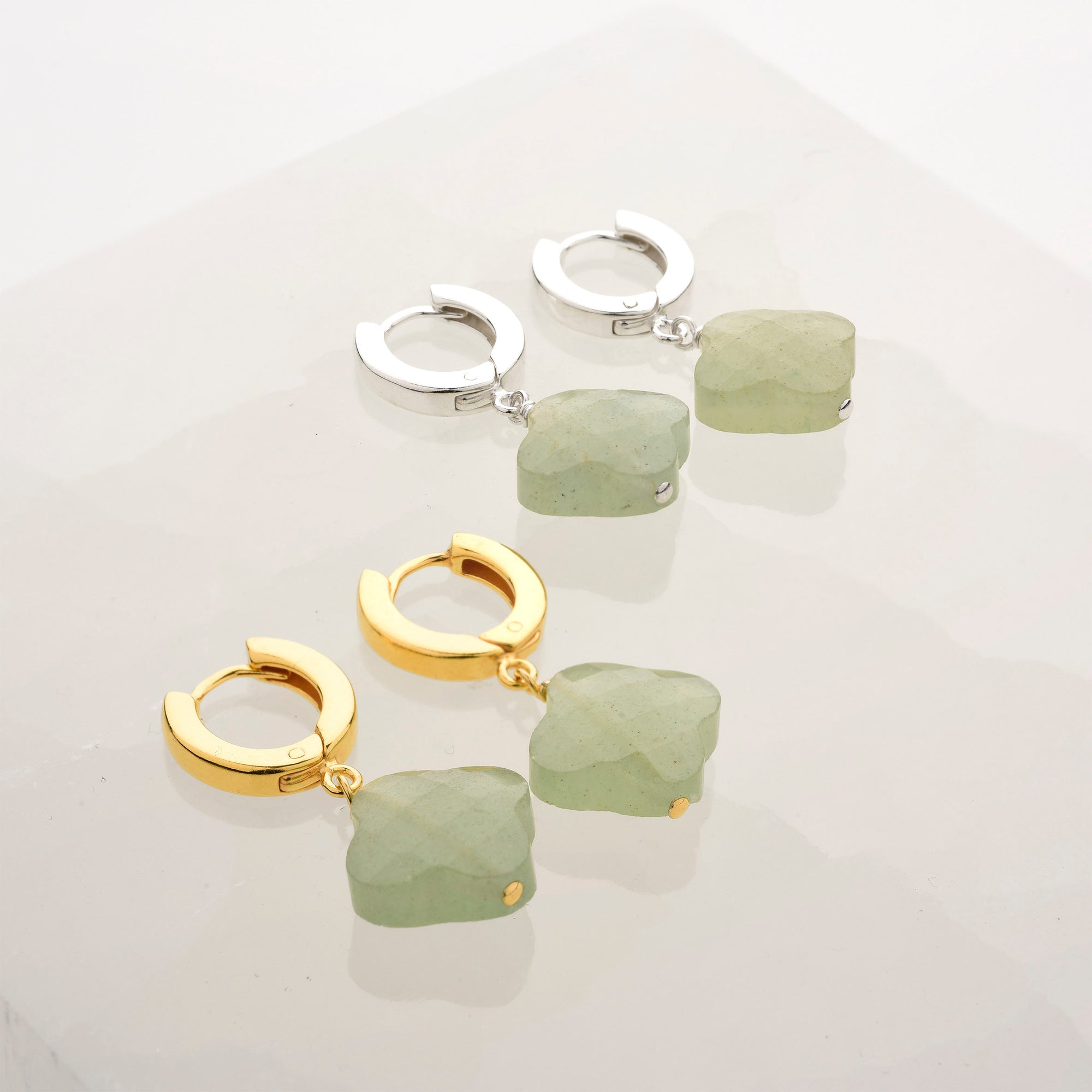 Two pairs of Clover Gem Hoop Earrings with green, faceted gemstone pendants rest on a glossy surface—one pair features gold finish hoops and the other sterling silver; both showcase similar stone shapes and size.