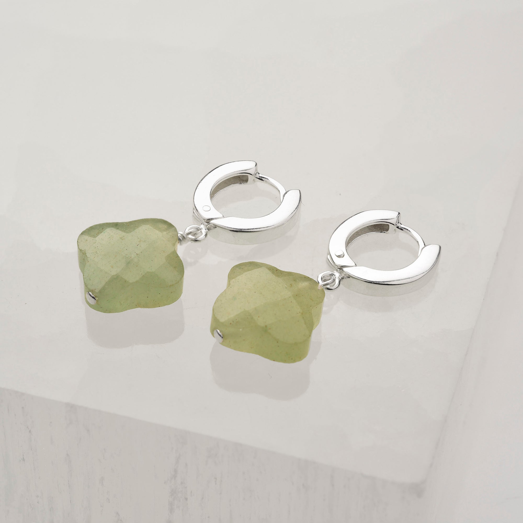The Clover Gem Hoop Earrings feature sterling silver hoops with green, faceted gemstone flower pendants, beautifully showcased on a white surface.