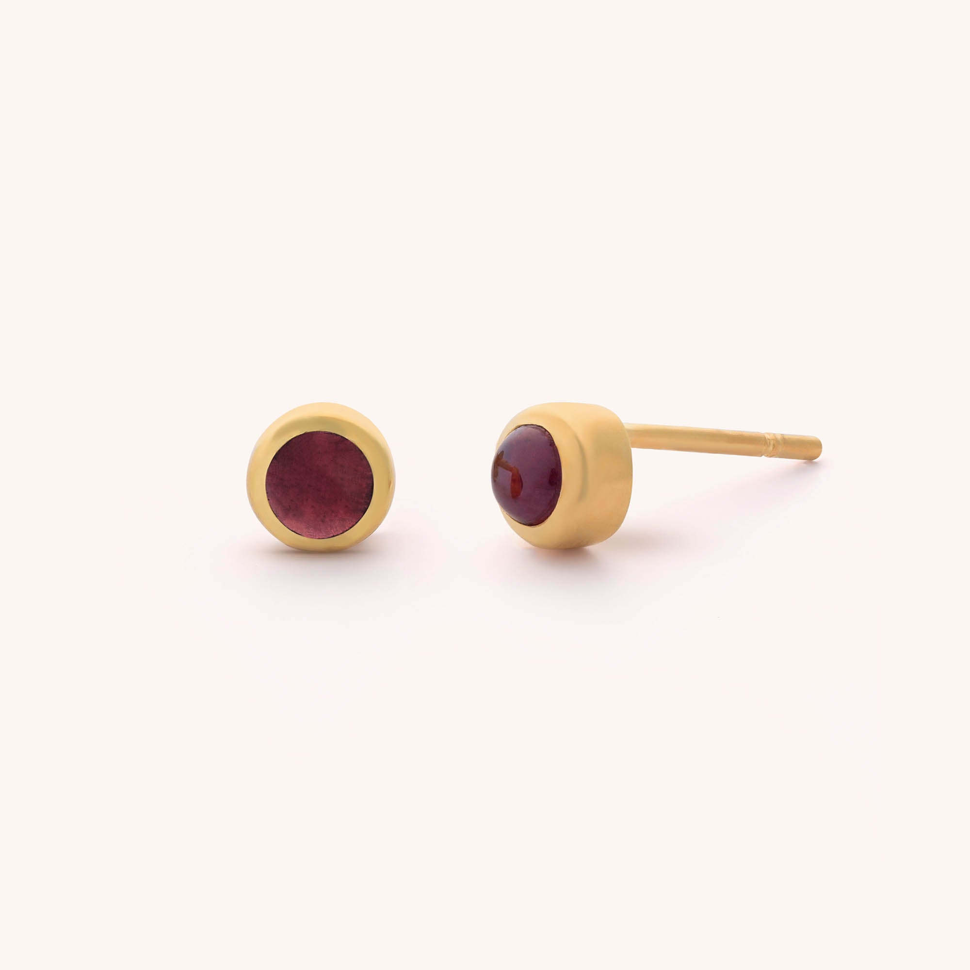 The Mini Gem Dot Birthstone Earrings feature gold studs with round, red gemstone cabochons. Displayed against a plain light background, one earring stands upright while the other shows its post and back, exuding elegance and charm.