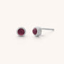 The Mini Gem Dot Birthstone Earrings feature two silver studs with round, dark red gemstone cabochons in the center, crafted from 925 sterling silver. Their classic appeal is enhanced against a plain light background.