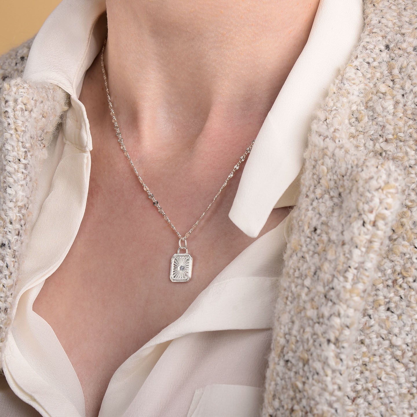 A person wears the Golden Dawn Necklace—a sterling silver chain with a rectangular, engraved sunburst pendant—styled with a cream blouse and textured beige cardigan.
