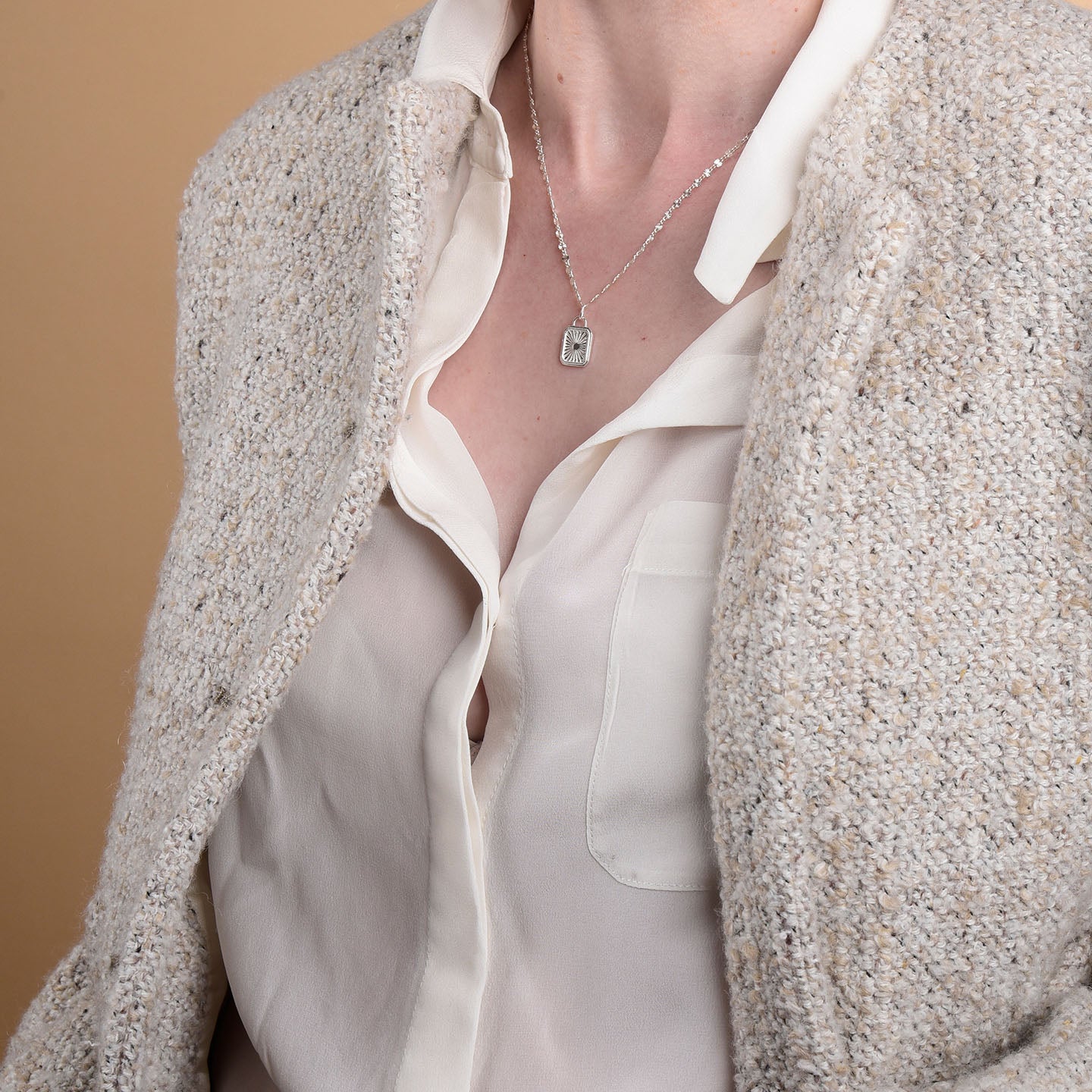 A person wears a cream textured jacket over an open white shirt, revealing the Golden Dawn Necklace with its rectangular pendant. The background is plain beige.