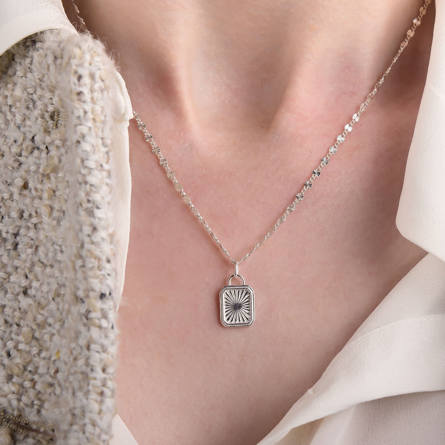 A person wears a textured cream jacket and white shirt, highlighting the Golden Dawn Necklace—a sterling silver chain with a sunburst pendant.