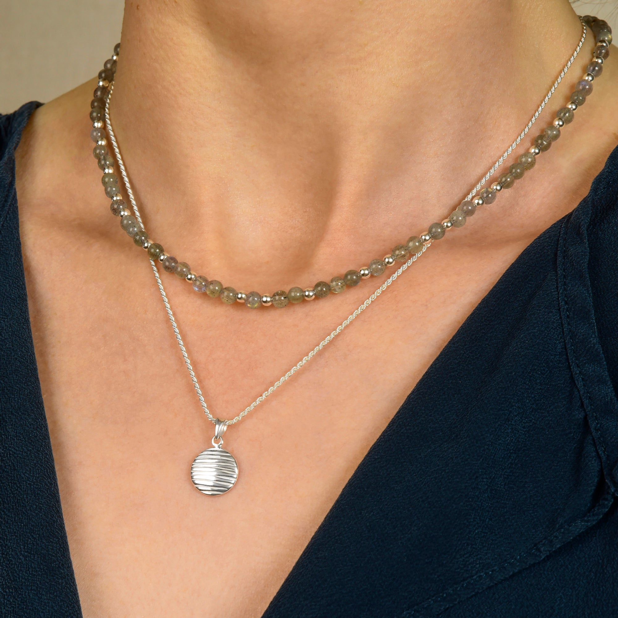 A person showcases modern elegance wearing a grayish-brown beaded choker and the Wave Disc Necklace on an adjustable rope chain, paired with a dark blue V-neck top.