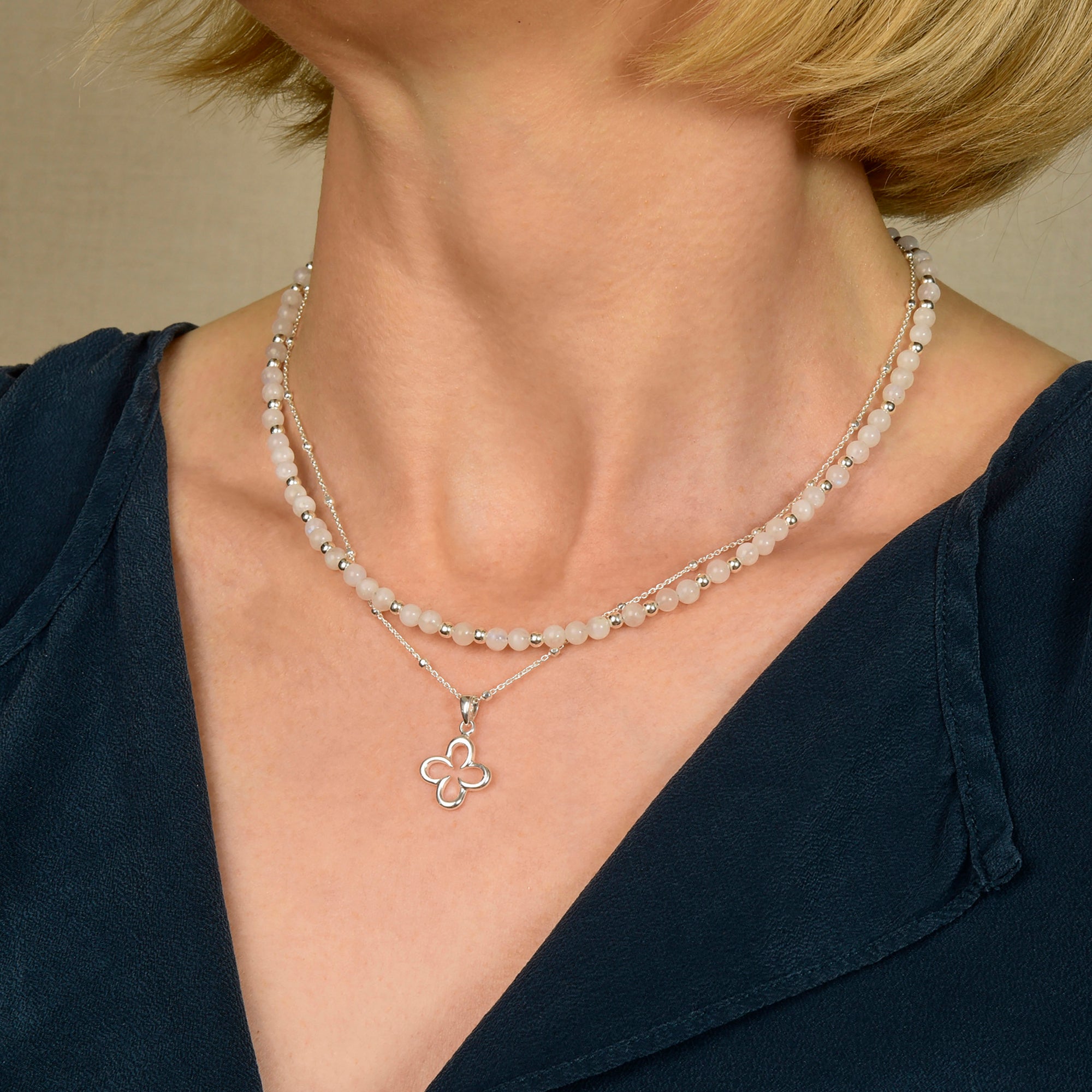 A woman in a navy V-neck top wears two necklaces: a beaded choker and the Jewel Bead Birthstone Necklace - June, featuring an eco-friendly sterling silver chain with a Rainbow Moonstone four-leaf flower pendant. Her short blonde hair frames her face.