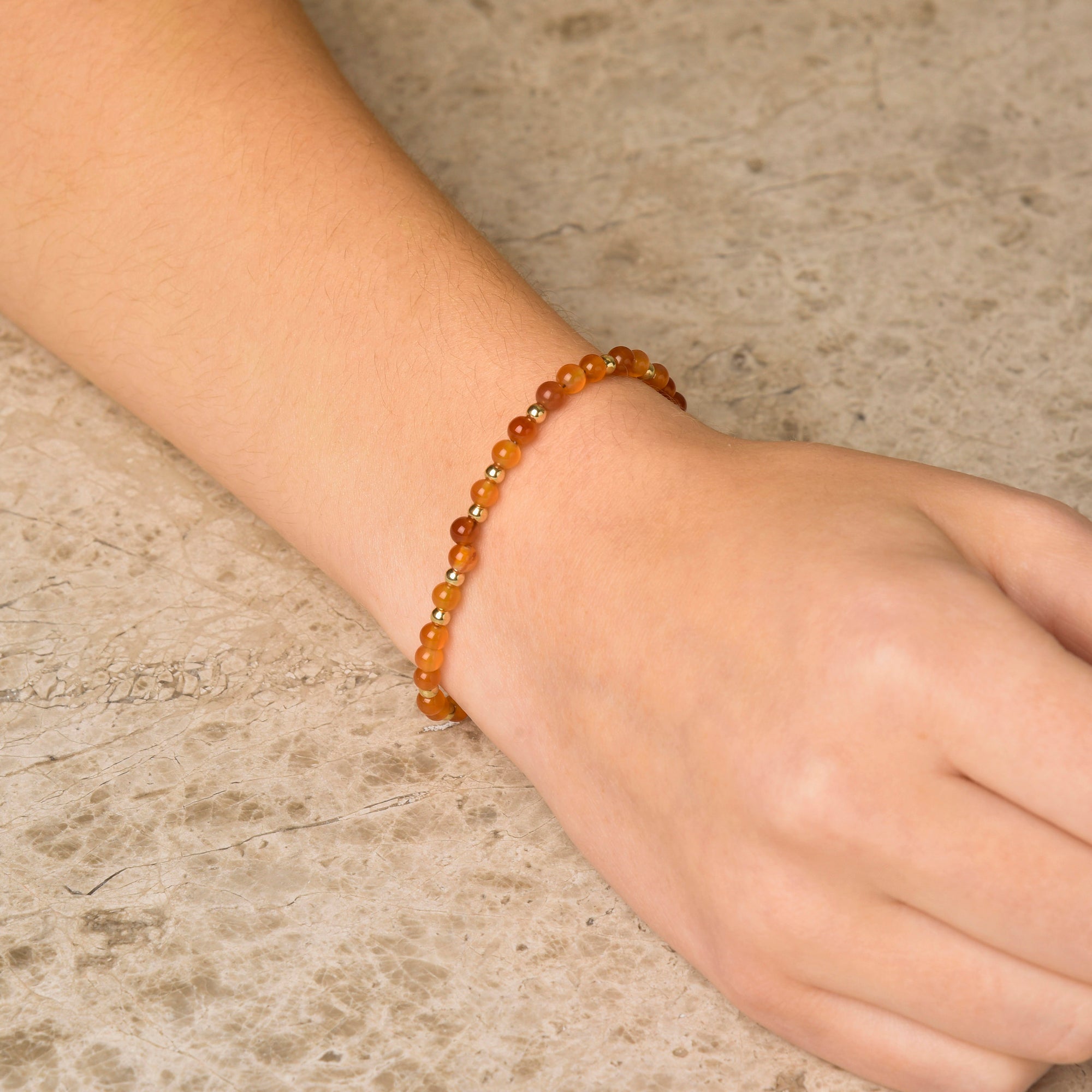 A hand rests on textured stone, wearing the Jewel Bead Bracelet—featuring small, round amber-colored natural gemstone beads.