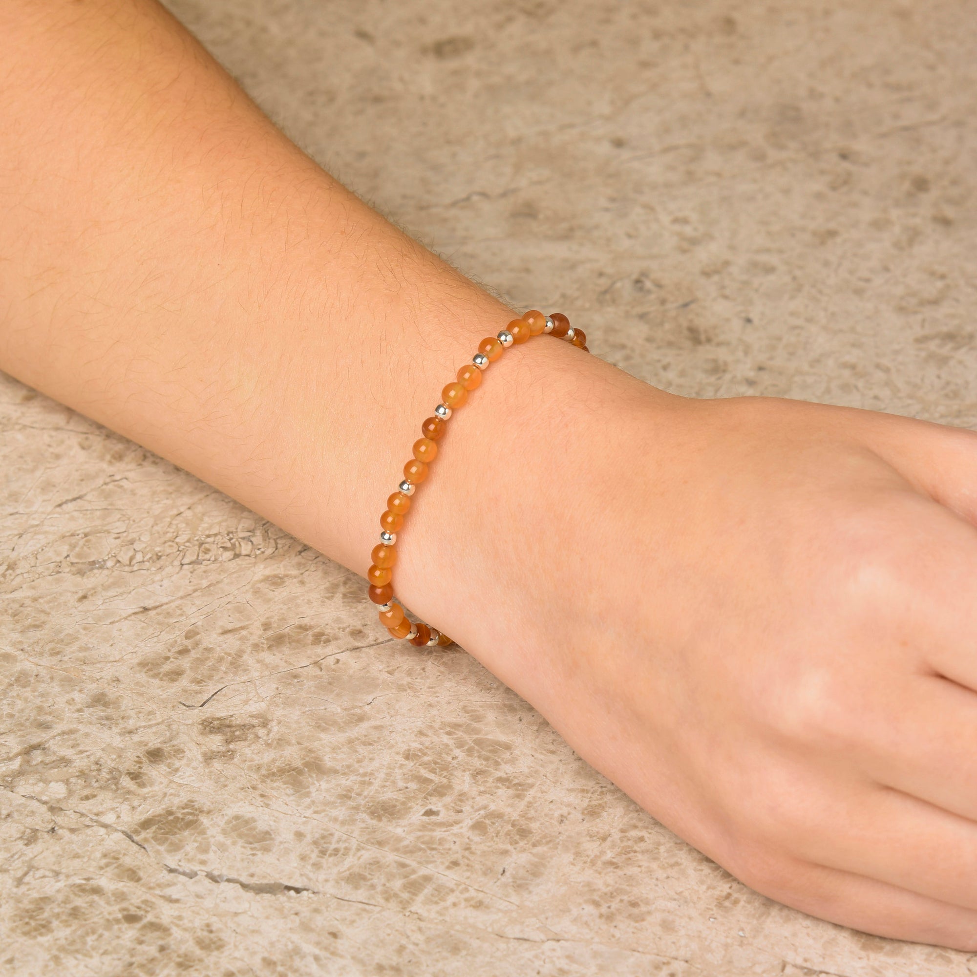 A hand and wrist wearing the Jewel Bead Bracelet, featuring small orange and eco-silver sterling silver beads, shown against a light stone background.