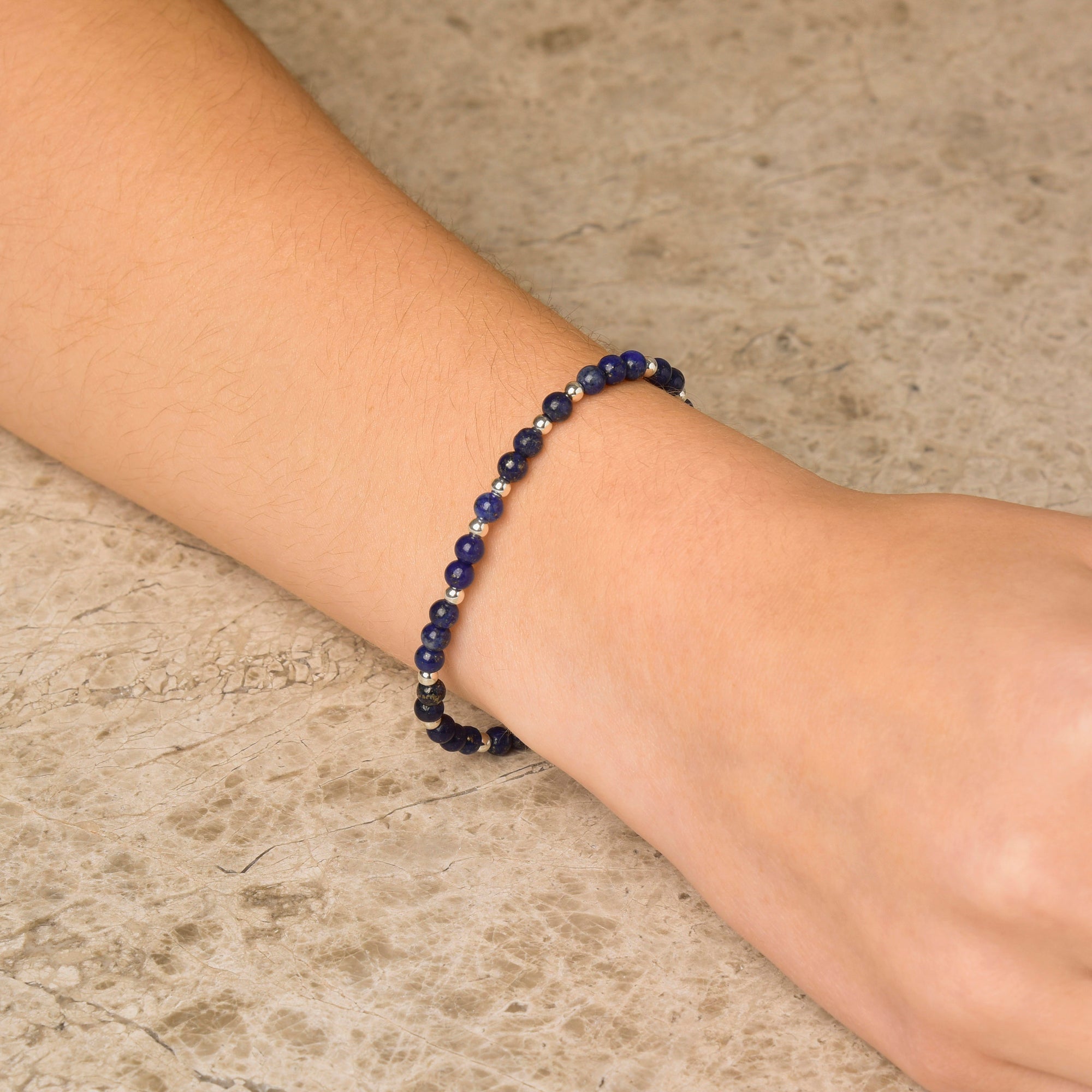 A close-up of a wrist wearing the Jewel Bead Bracelet, featuring small dark blue gemstones and eco-silver beads, displayed against a light stone surface.