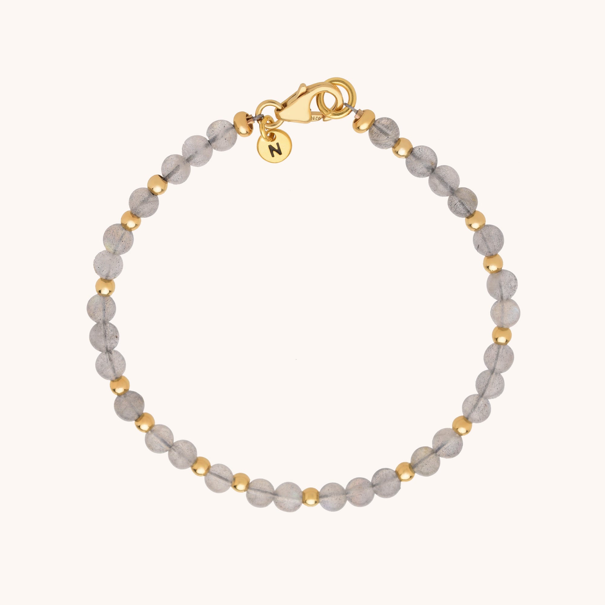 The Jewel Bead Bracelet features alternating small round grey and gold beads, a gold clasp, and a round gold charm engraved with the letter N, all displayed on a neutral background.