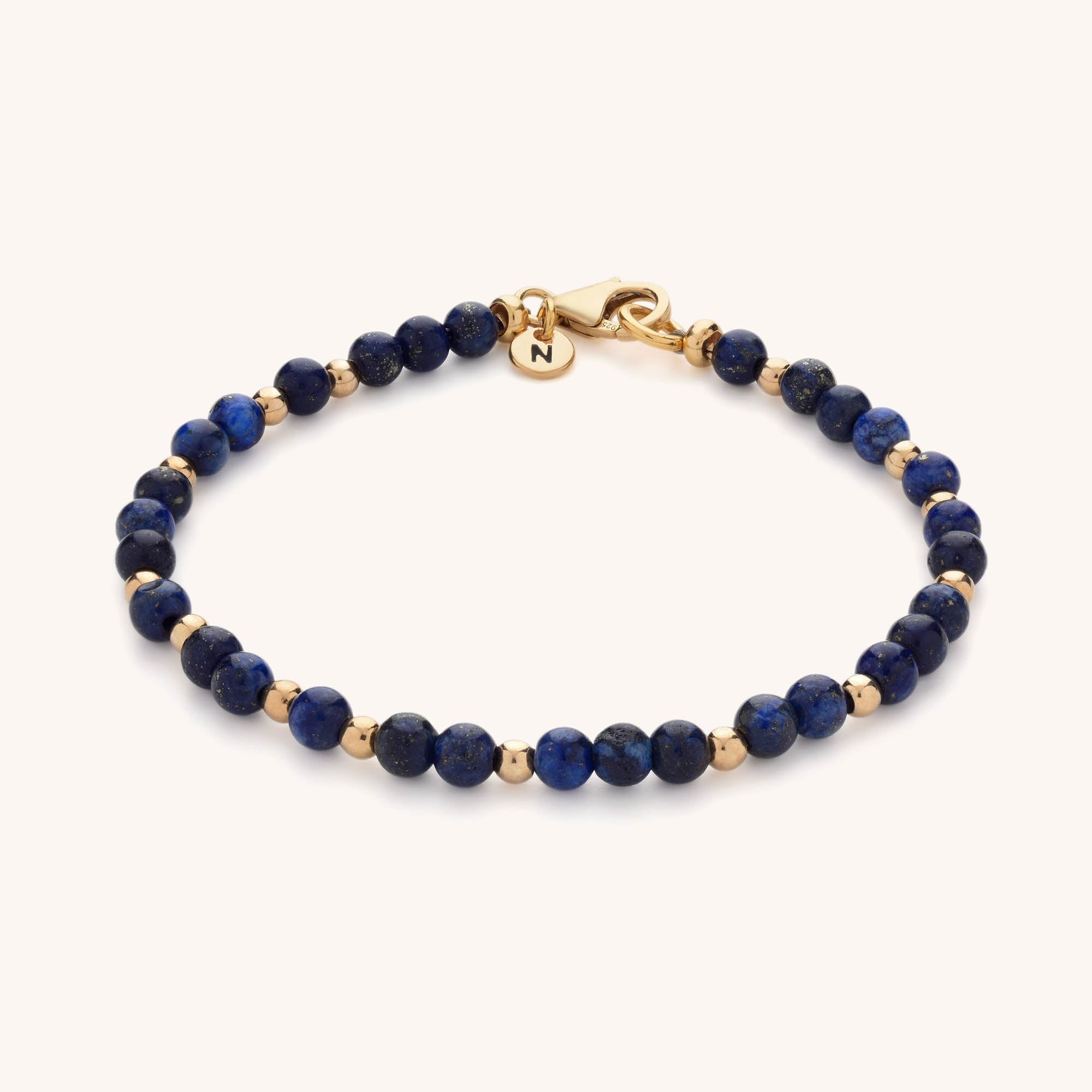 The Jewel Bead Birthstone Bracelet features alternating blue stones and gold beads, finished with a gold clasp and an engraved round gold charm with the letter N—ideal for handmade jewellery lovers.
