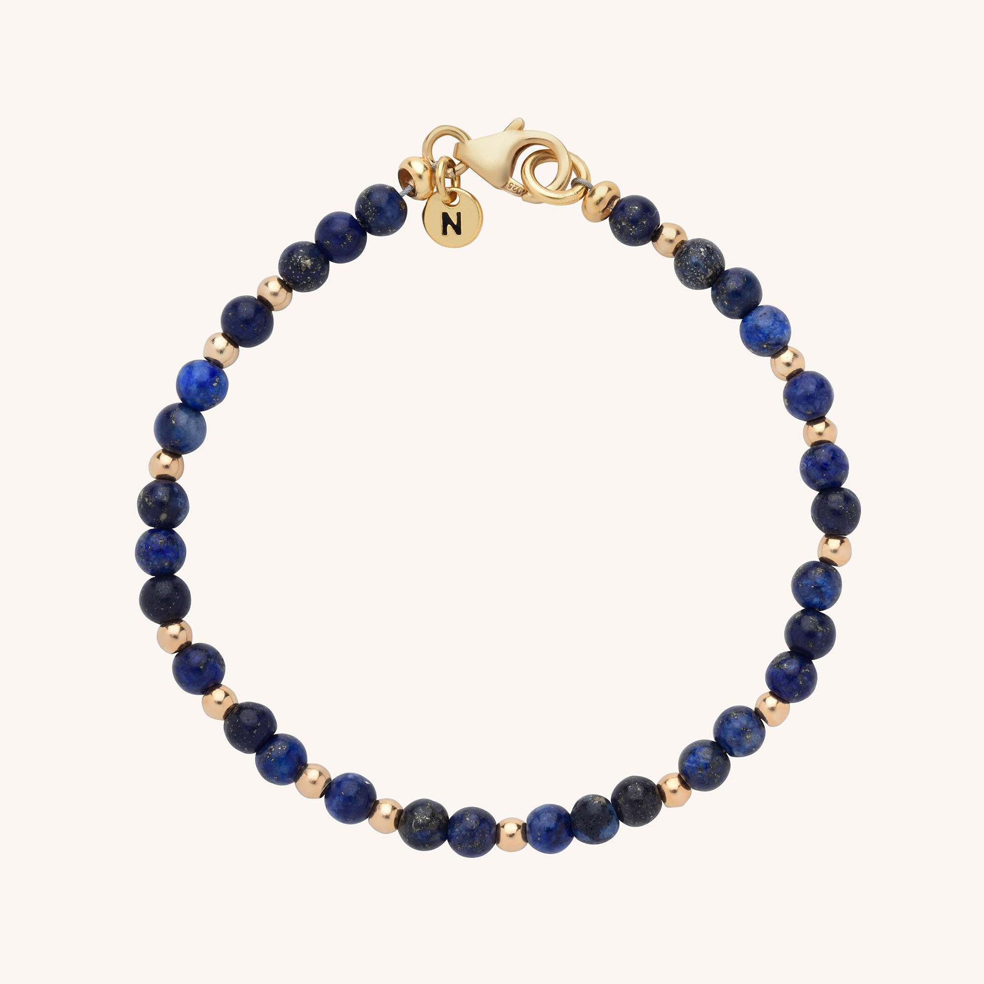 The Jewel Bead Birthstone Bracelet is a handmade piece featuring alternating dark blue and small gold beads, a gold lobster clasp, and a round gold charm engraved with the letter N.