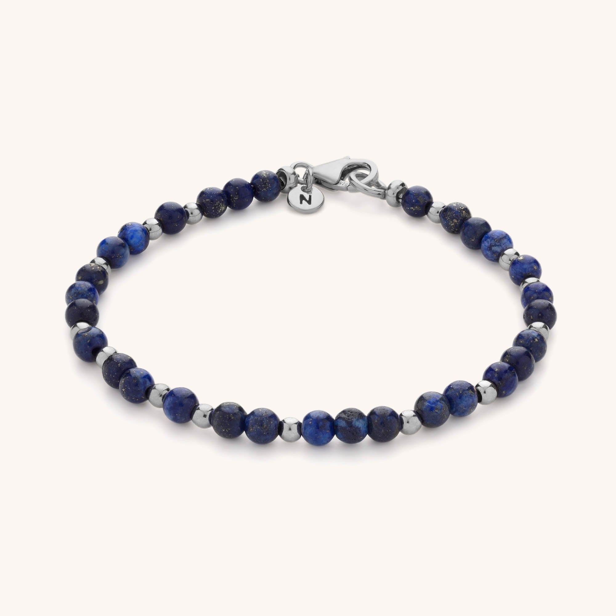 The Jewel Bead Birthstone Bracelet is handmade with round blue stones, silver spacer beads, a silver clasp, and a circular charm engraved with the letter N.