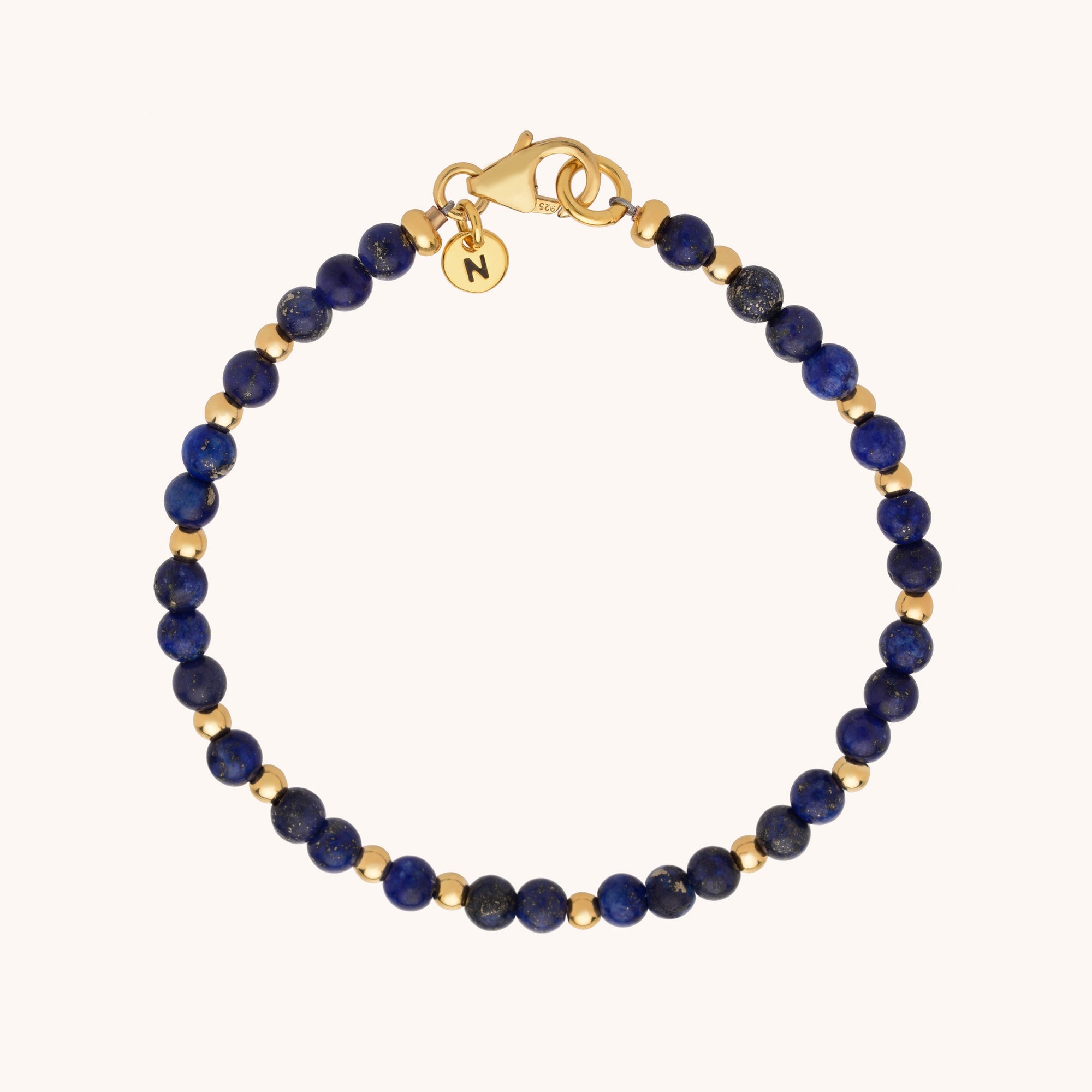 The Jewel Bead Bracelet showcases small round blue natural gemstones alternating with gold beads, finished with a gold clasp and a round charm engraved with the letter N.