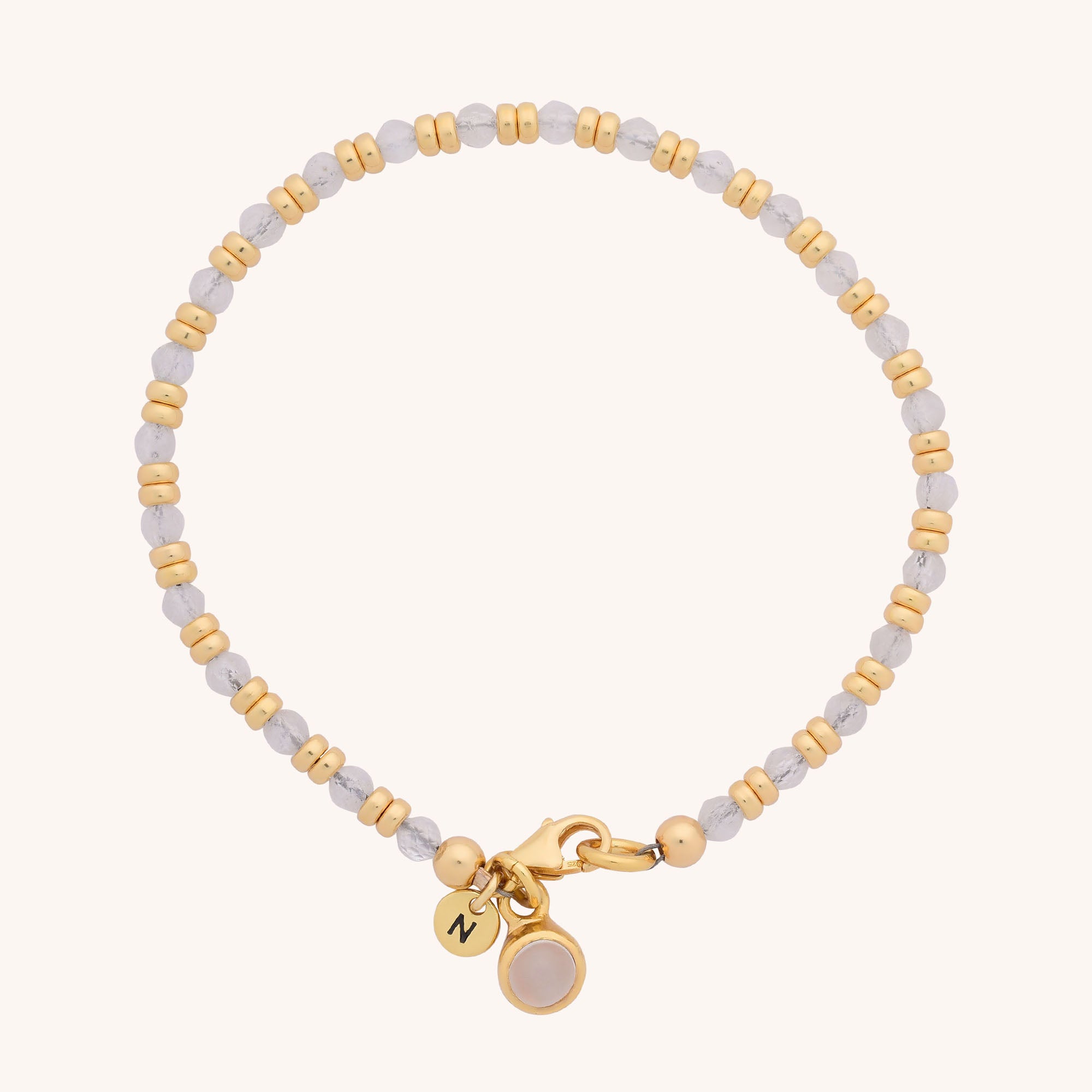 The Linear Birthstone Bracelet features alternating small gold and white beads, a gold clasp, and two charms—a letter N and a round, light-colored natural gemstone—perfect for fans of semi precious birthstone jewelry.