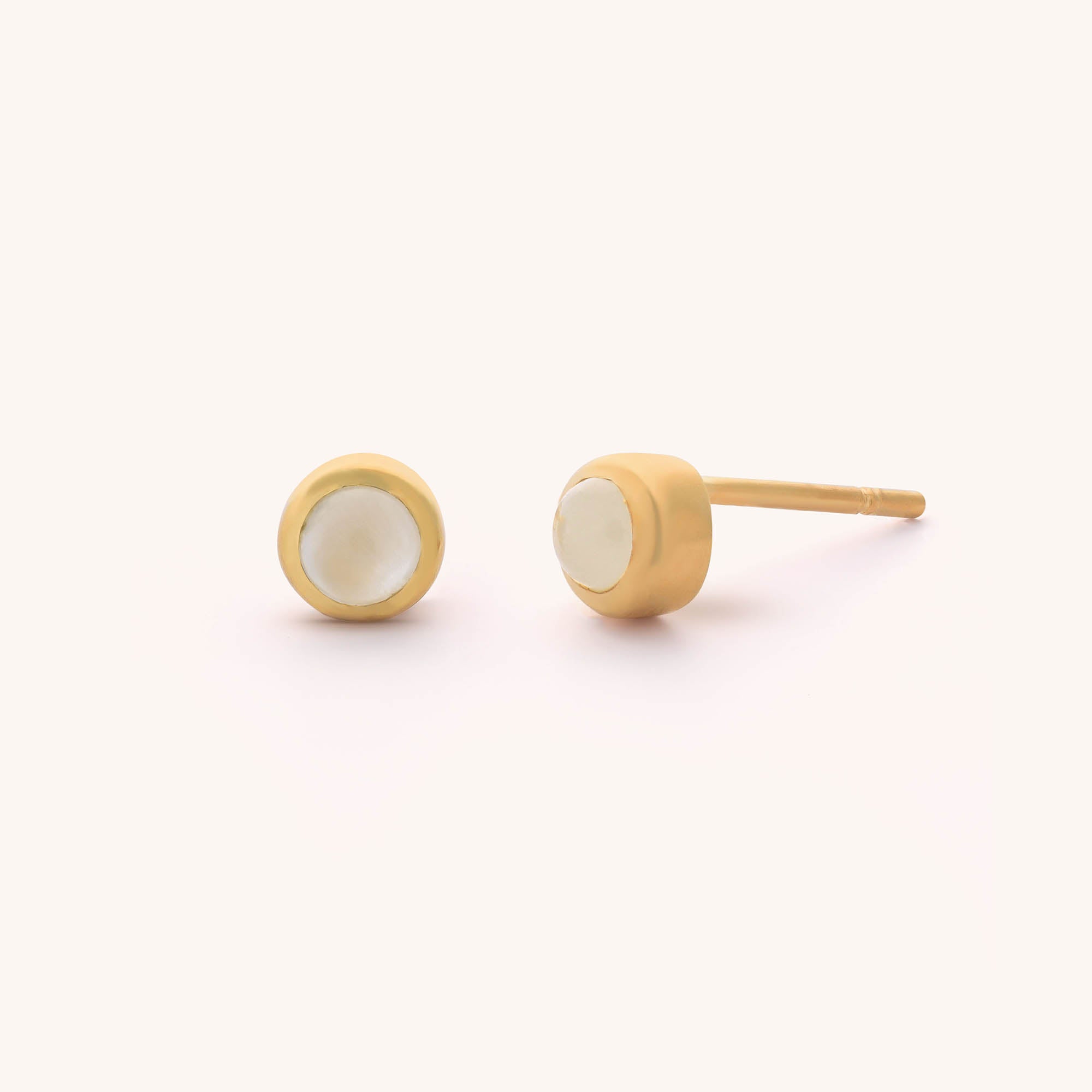 The Mini Gem Dot Birthstone Earrings - June feature round, smooth white moonstone centers. One gold earring lies flat to display the stone, while the other stands upright, revealing the post against a soft off-white background.