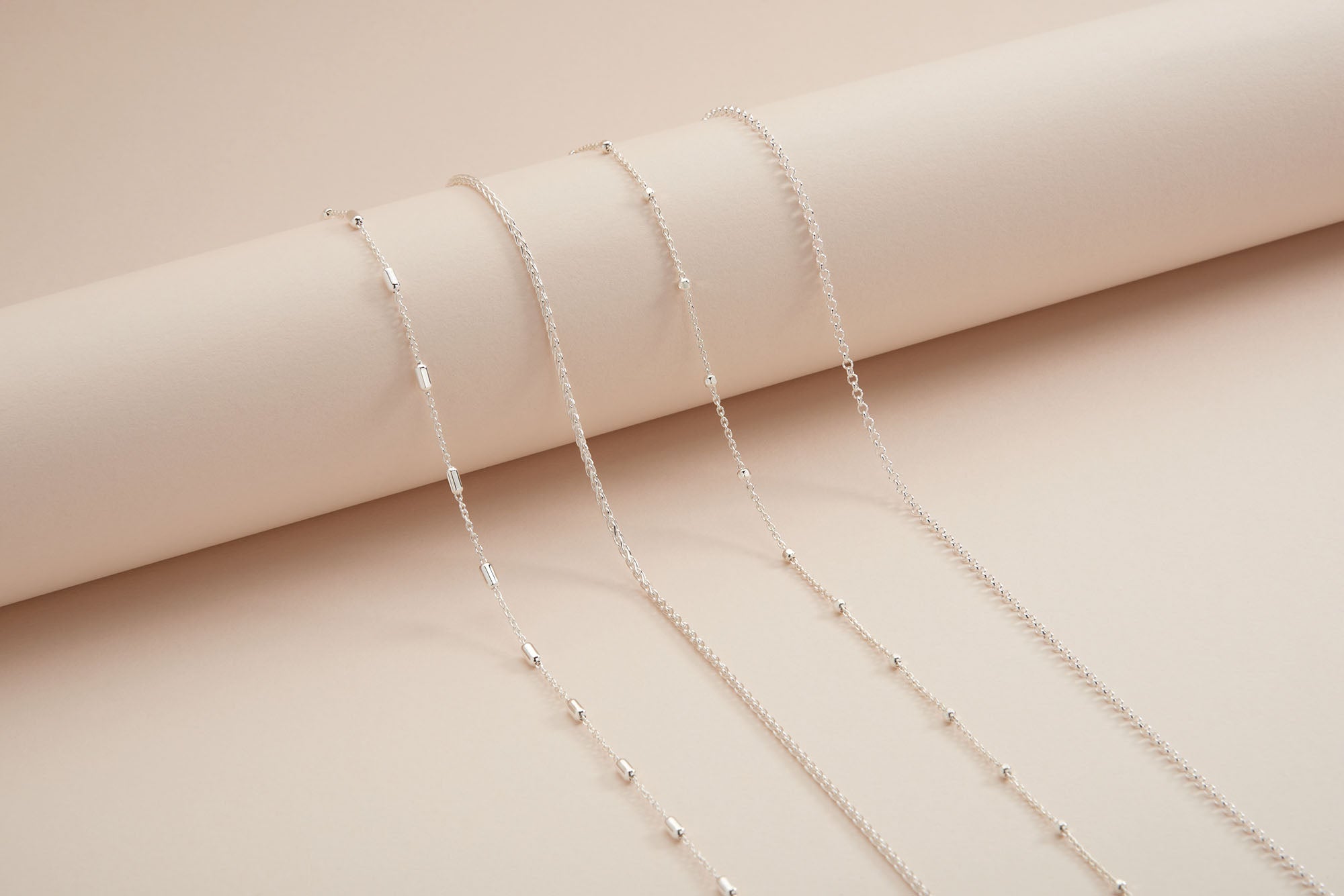 Four delicate silver necklaces with different chain patterns are draped over a cream-colored cylindrical surface, displayed on a matching soft beige background.